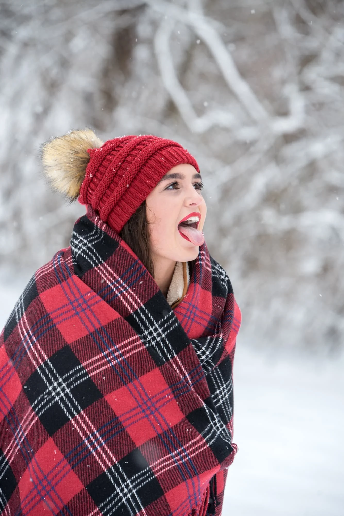 Whimsical winter senior photo of a girl wrapped in a red plaid blanket and red beanie, sticking her tongue out to catch falling snowflakes during a winter storm.