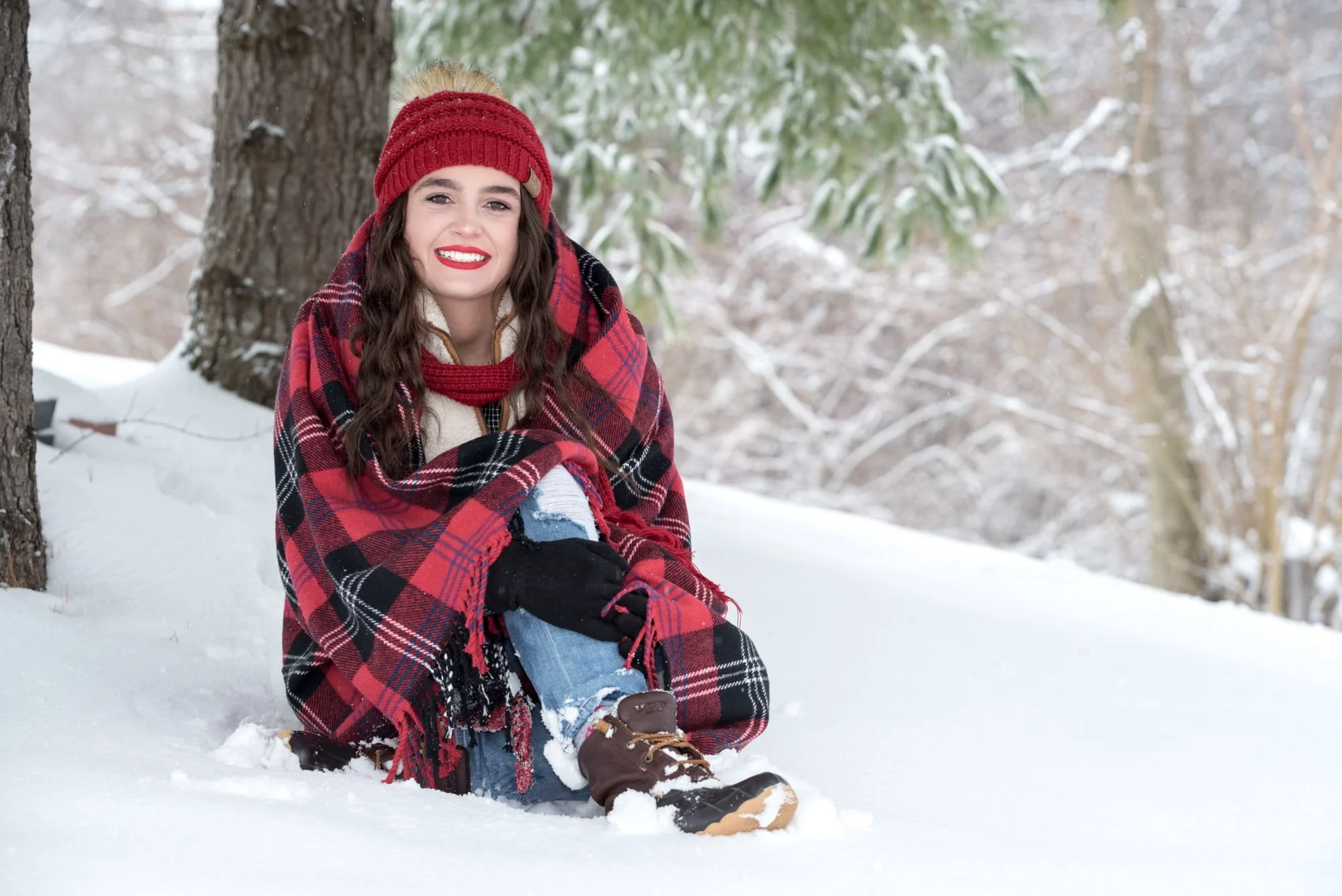 High school senior girl wrapped in a red plaid blanket, sitting in the snow under a tree wearing a red beanie, white vest, ripped jeans, and brown snow boots for a cozy winter portrait in Columbia IL.