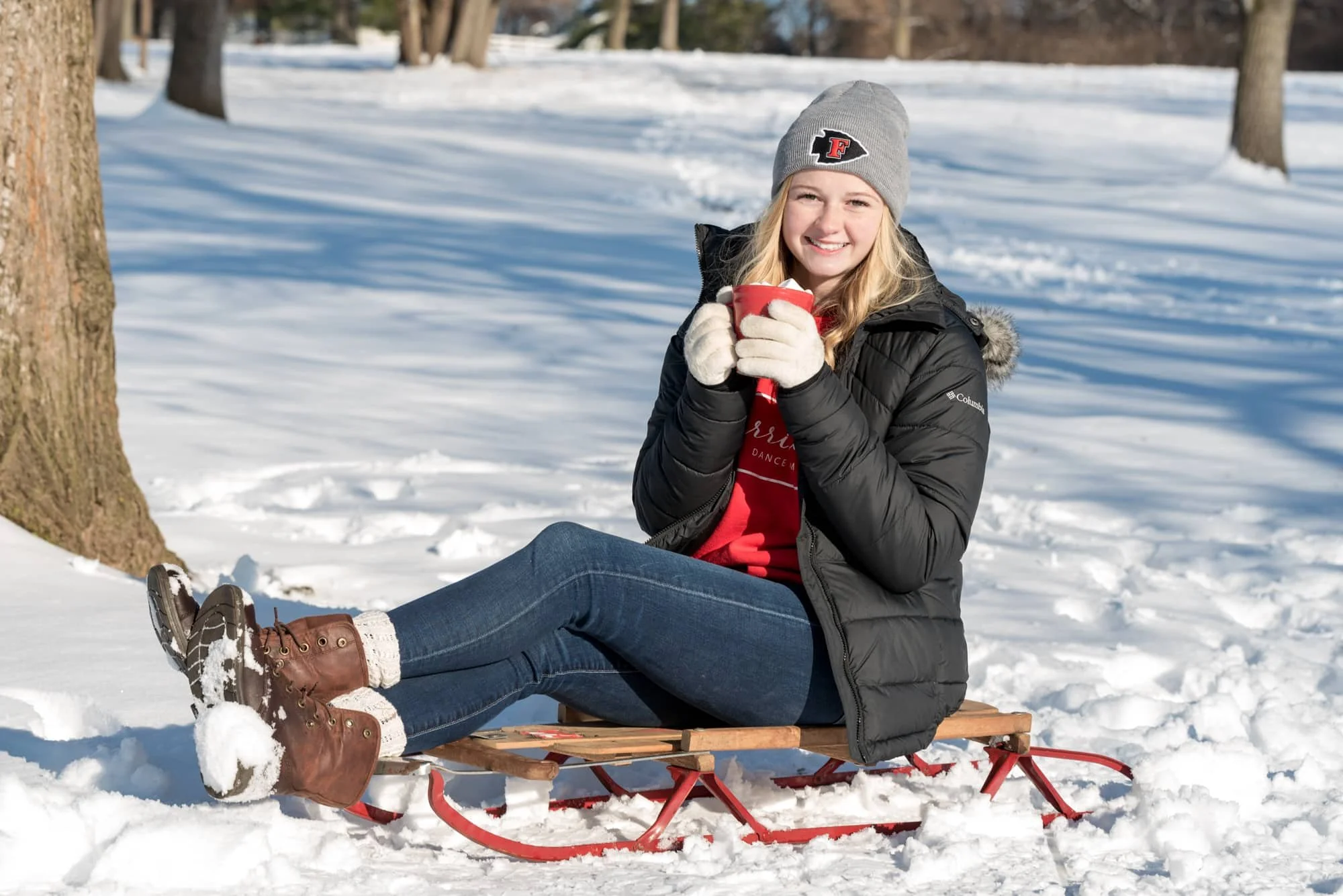 Fox High School senior girl smiling on a sled in the snow, wearing a grey Fox beanie, Fox Warriorettes red sweater, black coat, and winter boots while holding a red mug of hot chocolate with giant marshmallows at Suson Park in Arnold, MO.