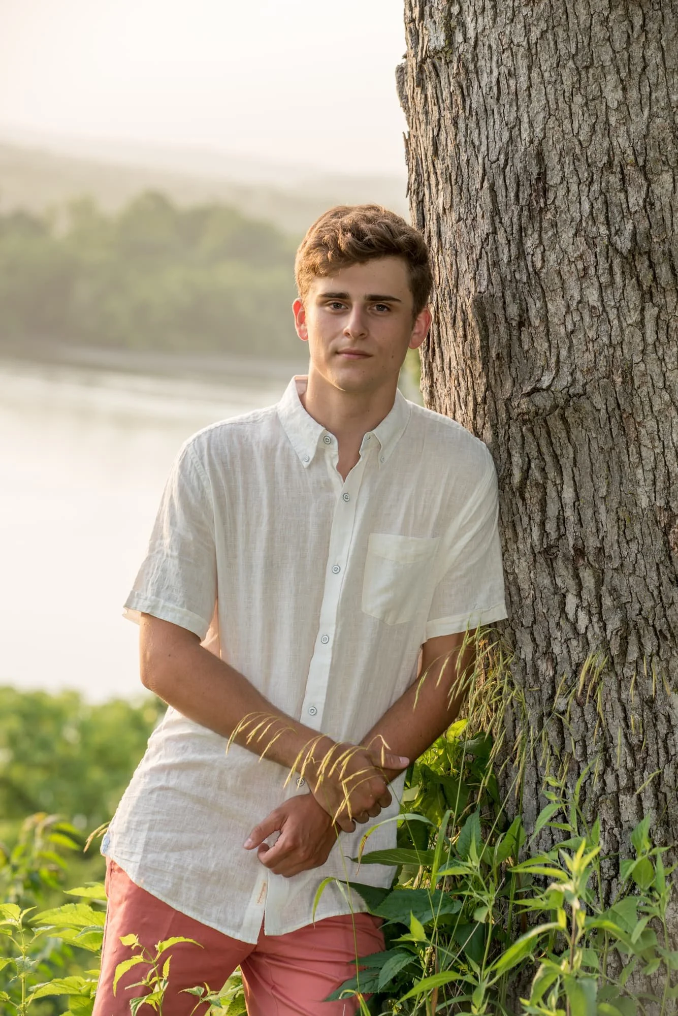 Senior guy wearing a white button-up shirt and coral shorts, leaning against a tree in the weeds in front of the river during a hazy Sahara desert dust sunset for a photo in Oakville MO.