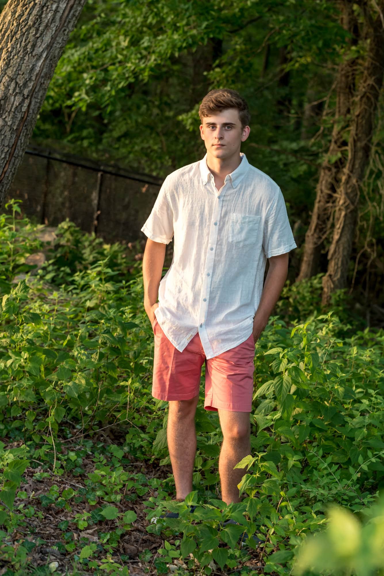 Senior-Boy-Senior guy with a serious expression wearing a white button-up shirt and coral shorts, standing in the weeds during a sunset session in Oakville MO.-In-Woods