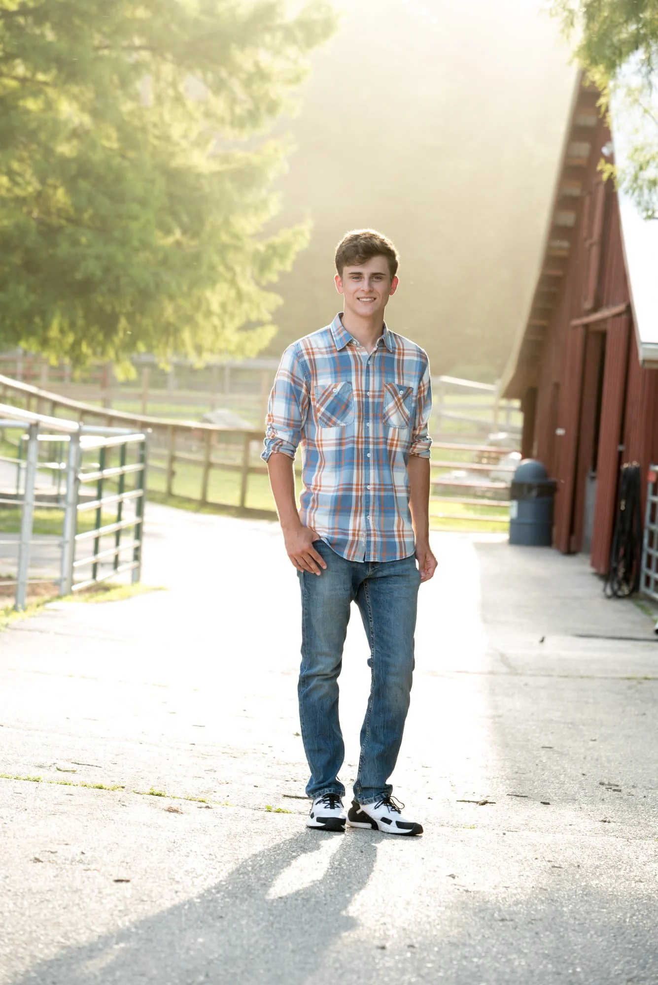 Senior picture of a guy wearing a blue and orange plaid shirt, blue jeans, and white tennis shoes, standing on a pathway in front of a red barn during a sunset illuminated by hazy Sahara desert dust at Suson Park.