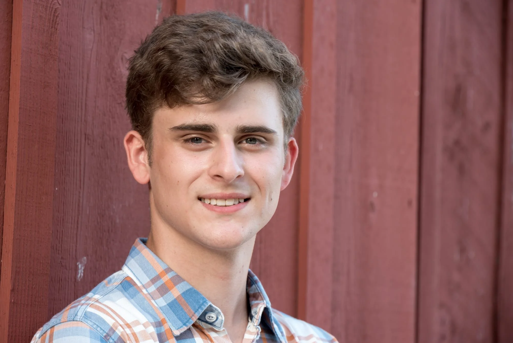 Close-up senior picture of a guy wearing a blue and orange plaid shirt, standing in front of a red barn at Suson Park.