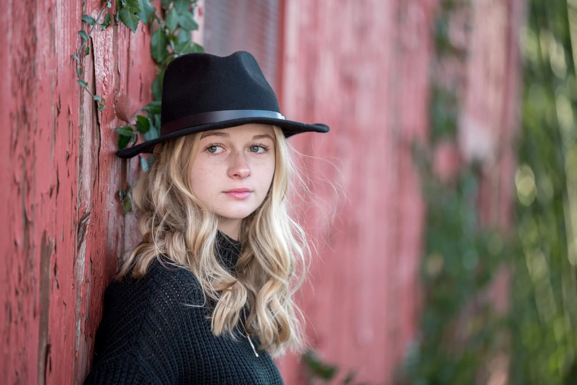 Moody autumn senior picture taken by a Jefferson County senior photographer, featuring a girl wearing a black Fedora and black sweater, leaning back against a weathered red barn covered in ivy while looking away with a serious expression.