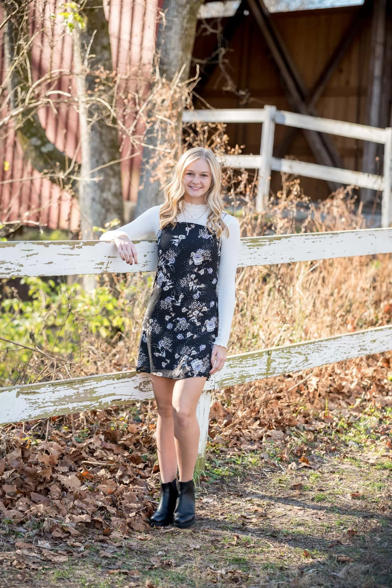 Picturesque outdoor senior picture featuring a girl resting her arm on a weathered white wooden fence, with the iconic red Sandy Creek Covered Bridge and peak autumn trees beautifully framing the background.