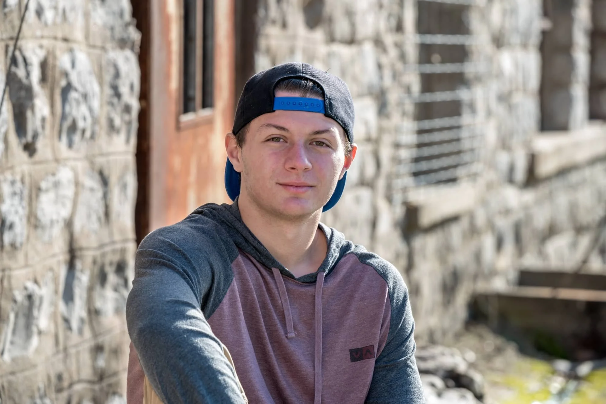 Senior guy wearing a backwards baseball cap and a grey and red hoodie, sitting in front of a weathered stone building and a rusted metal door for a rugged industrial portrait at Rock City in Valmeyer IL.