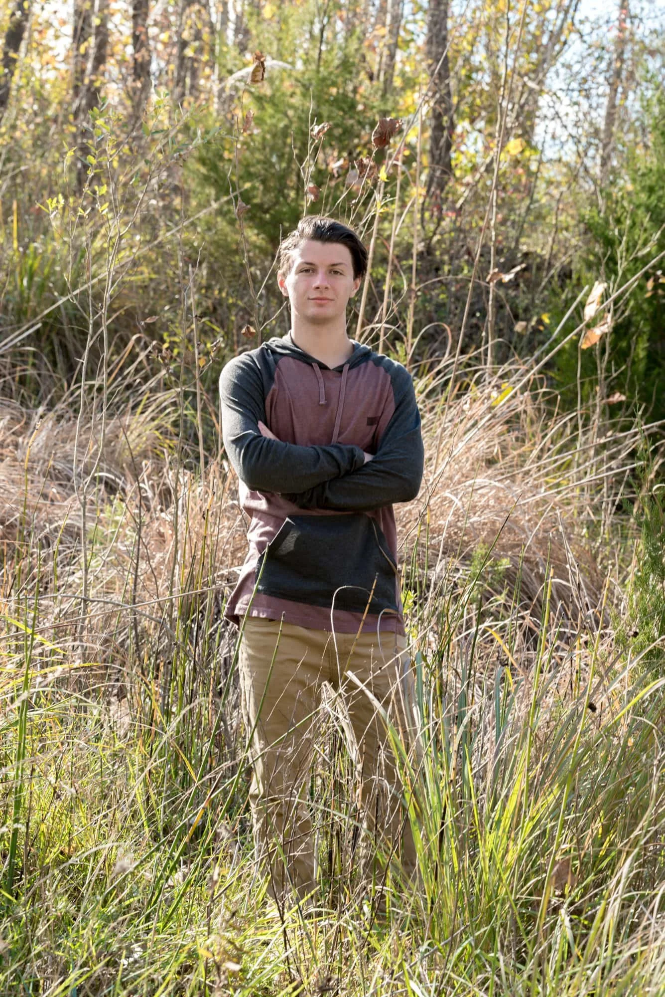Senior photo of a guy with a serious face, standing with his arms crossed in a field of tall grassy weeds, wearing a grey and red hoodie with khaki pants for a rustic country session in Valmeyer IL.