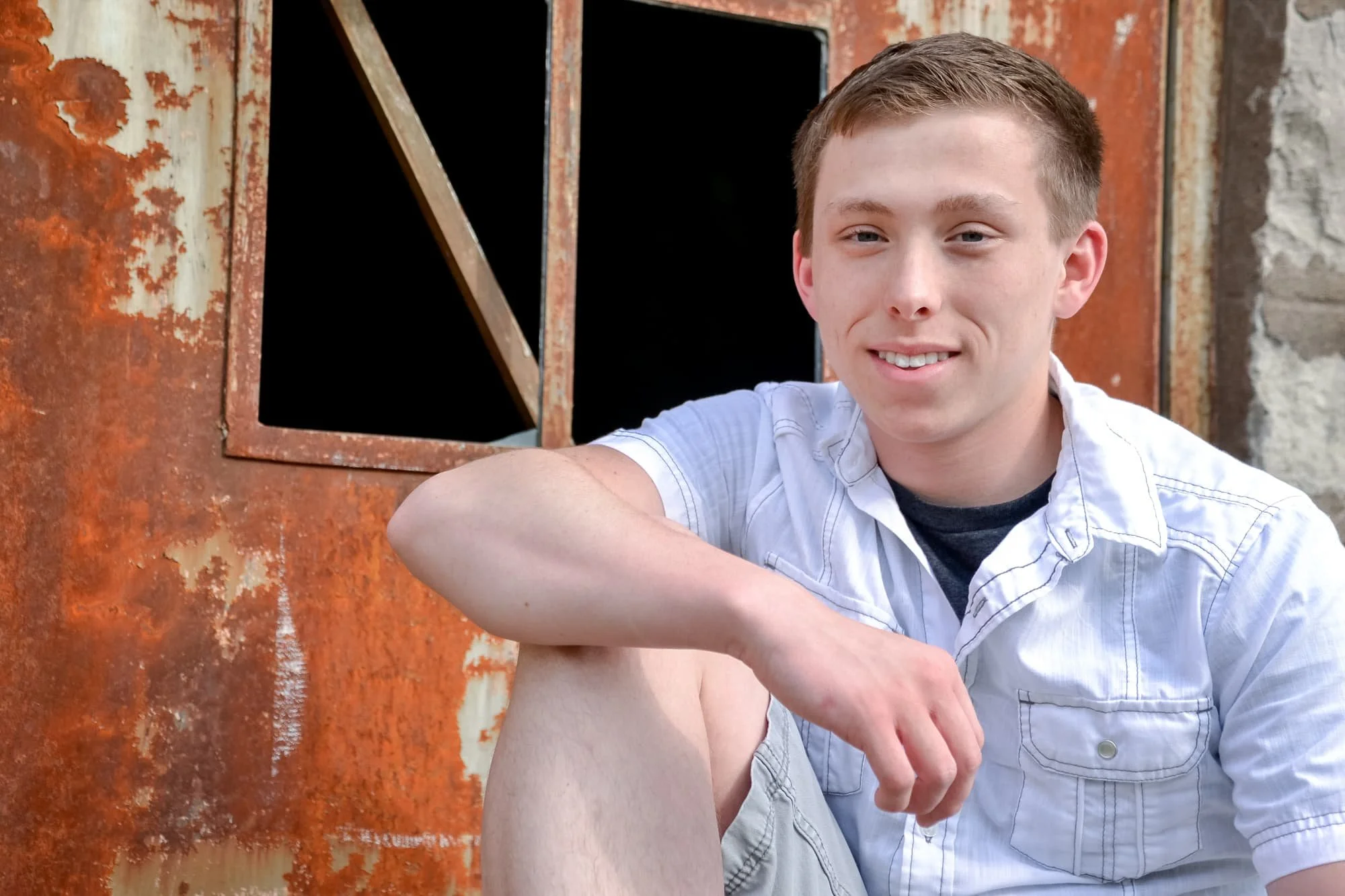 Senior guy wearing a white shirt, sitting with his arm on his knee in front of a rustic stone building and an old rusty metal door for a unique industrial senior photo at Rock City in Valmeyer IL.