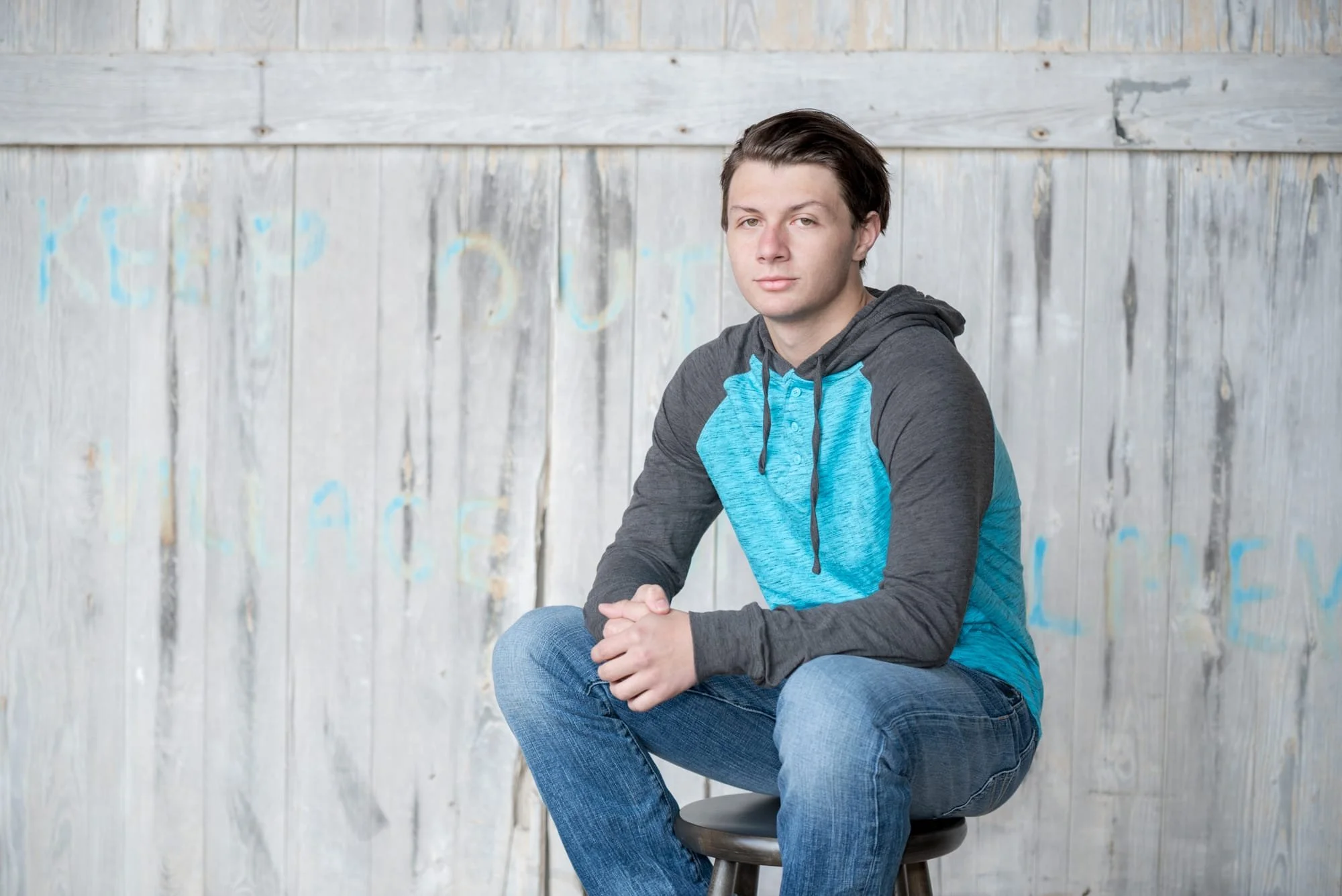 Senior picture of a guy wearing a grey and blue hoodie with blue jeans, sitting on a stool with his hands together in front of a weathered grey wooden door for a rustic portrait at Rock City in Valmeyer IL.