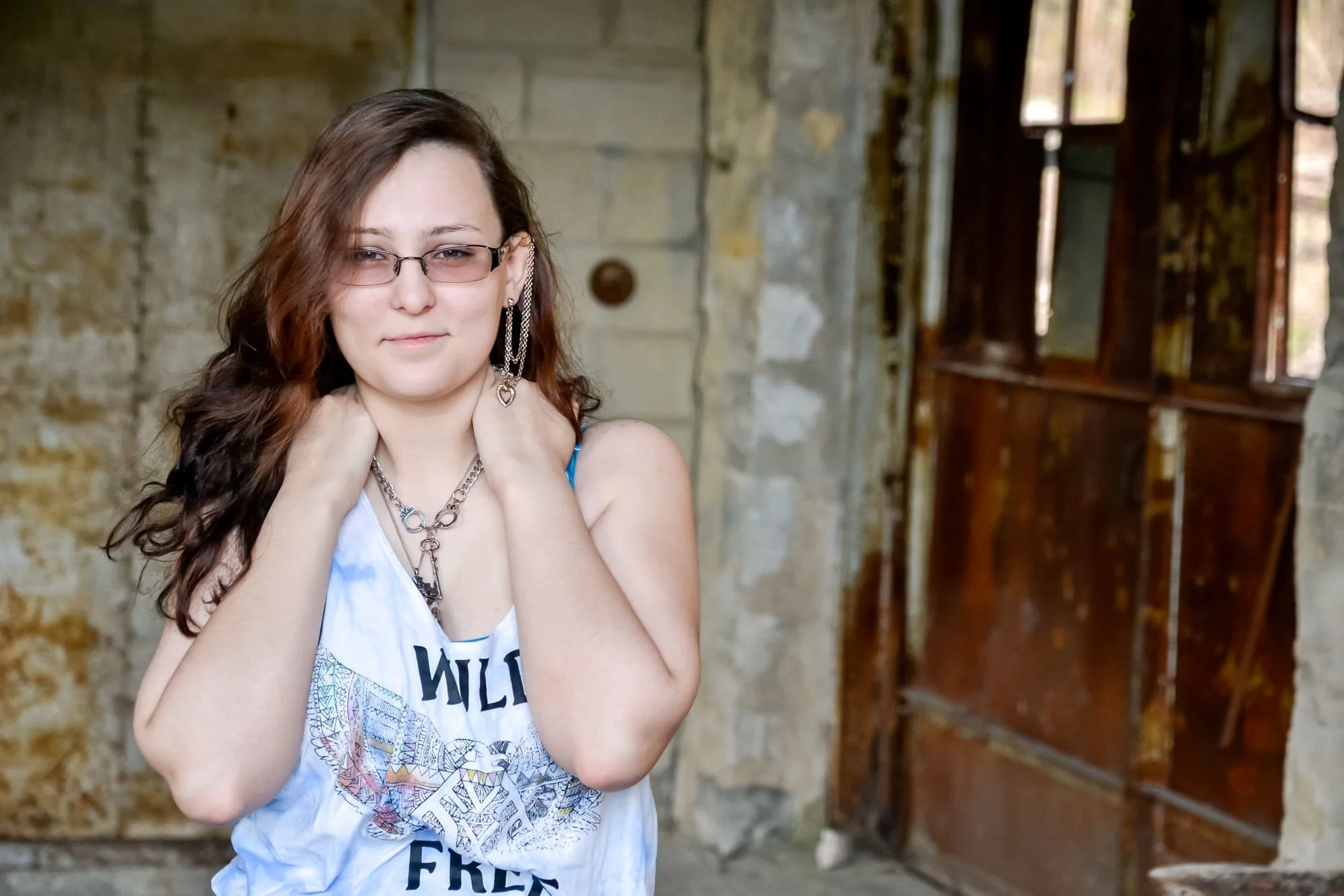 Close-up senior photo of a girl wearing a white graphic tank top, a chunky necklace, and big earrings, posing with a soft smile in front of a rustic stone wall and a weathered, rusty door at Rock City in Valmeyer IL.
