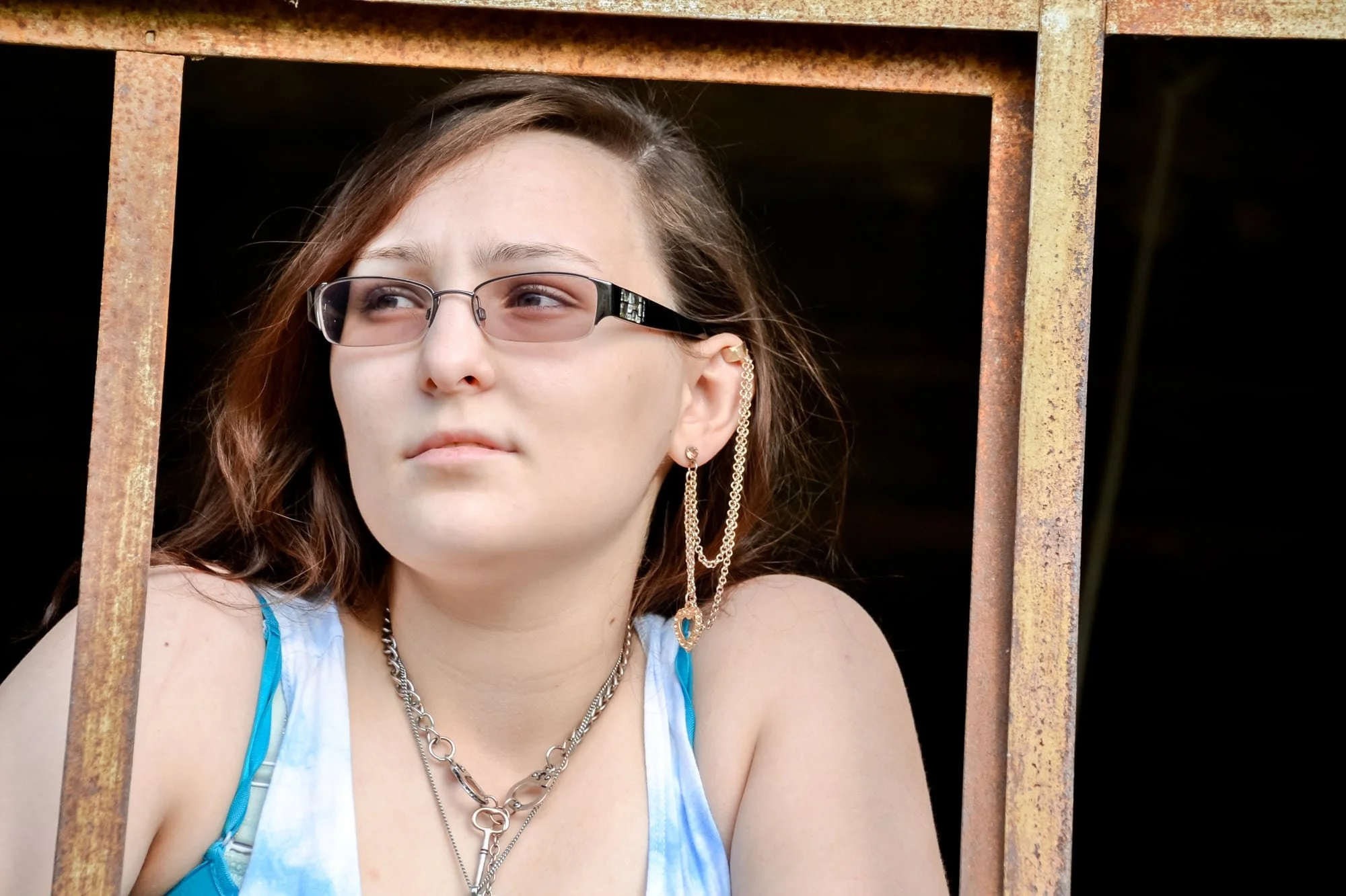 Close-up senior portrait of a girl wearing a chunky necklace and big earrings, looking away thoughtfully while peering through a rusted, glassless window frame at Rock City in Valmeyer IL.