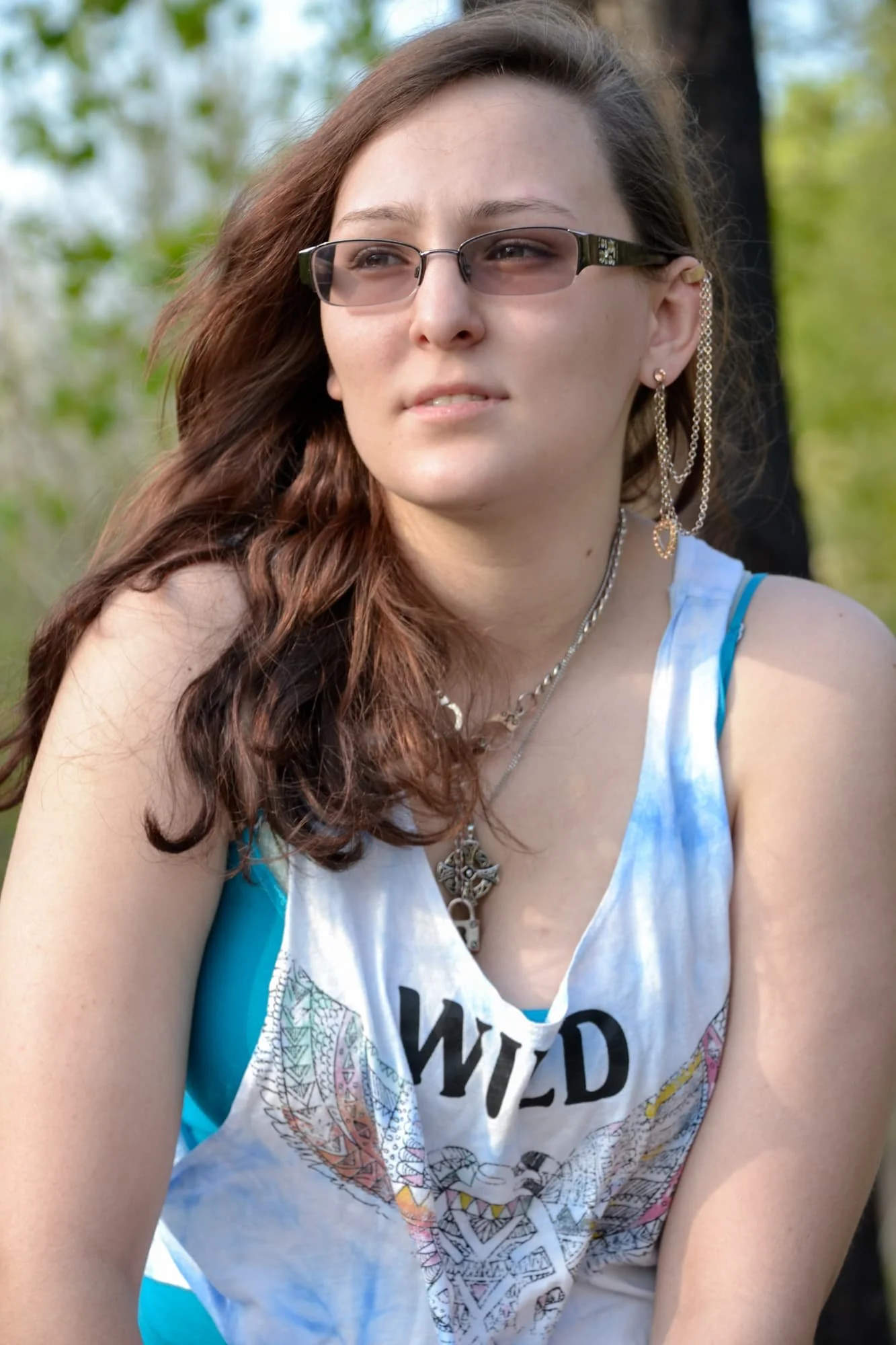 Senior girl with her hair blowing in the wind, wearing a white graphic tank top and a statement necklace while looking off into the distance in front of lush green foliage at Rock City in Valmeyer IL.