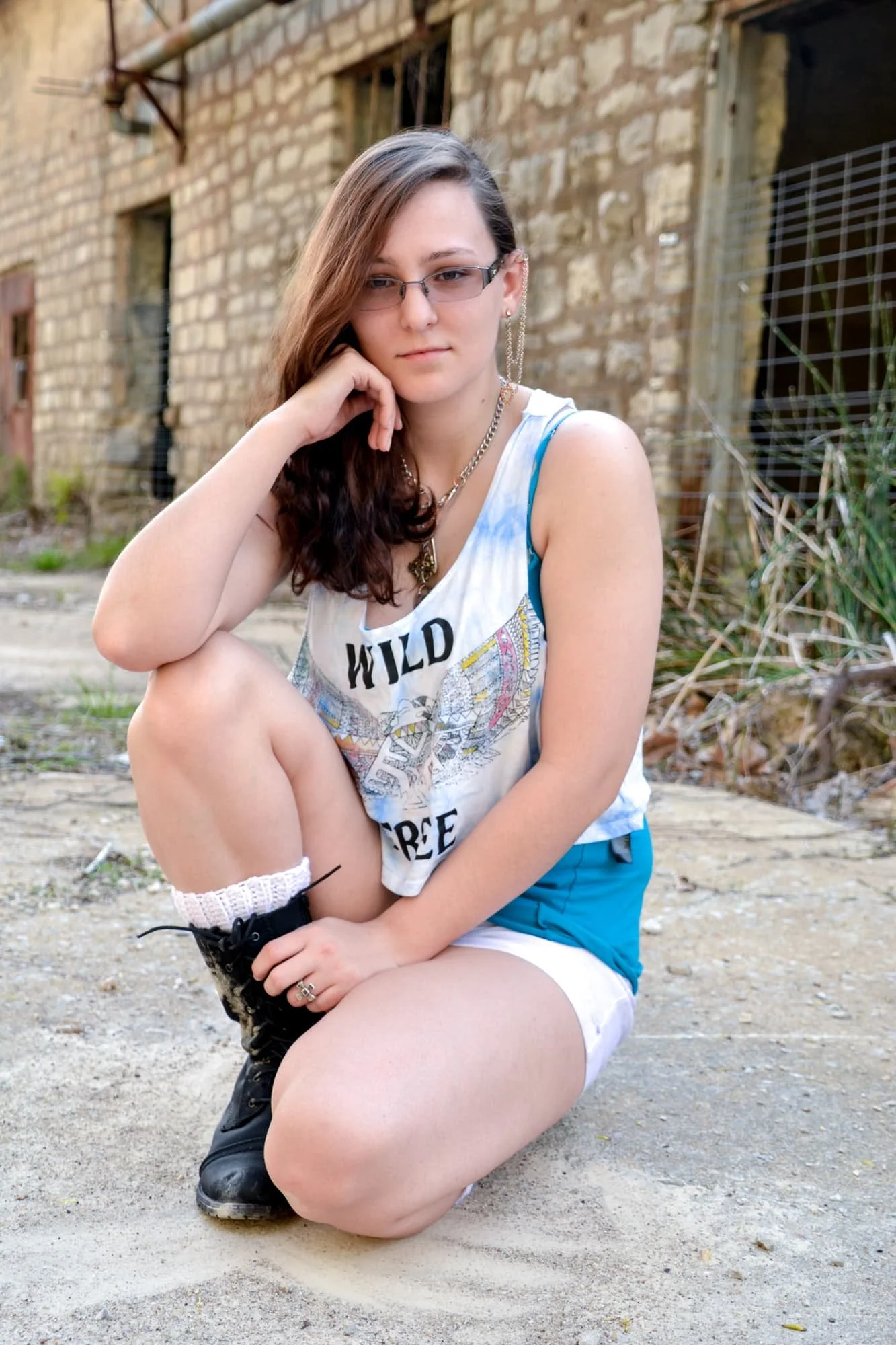 Senior picture of a girl crouched down with her elbow on her knee and her hand on her face, posing in front of an abandoned stone building with rusted metal elements at Rock City in Valmeyer IL.