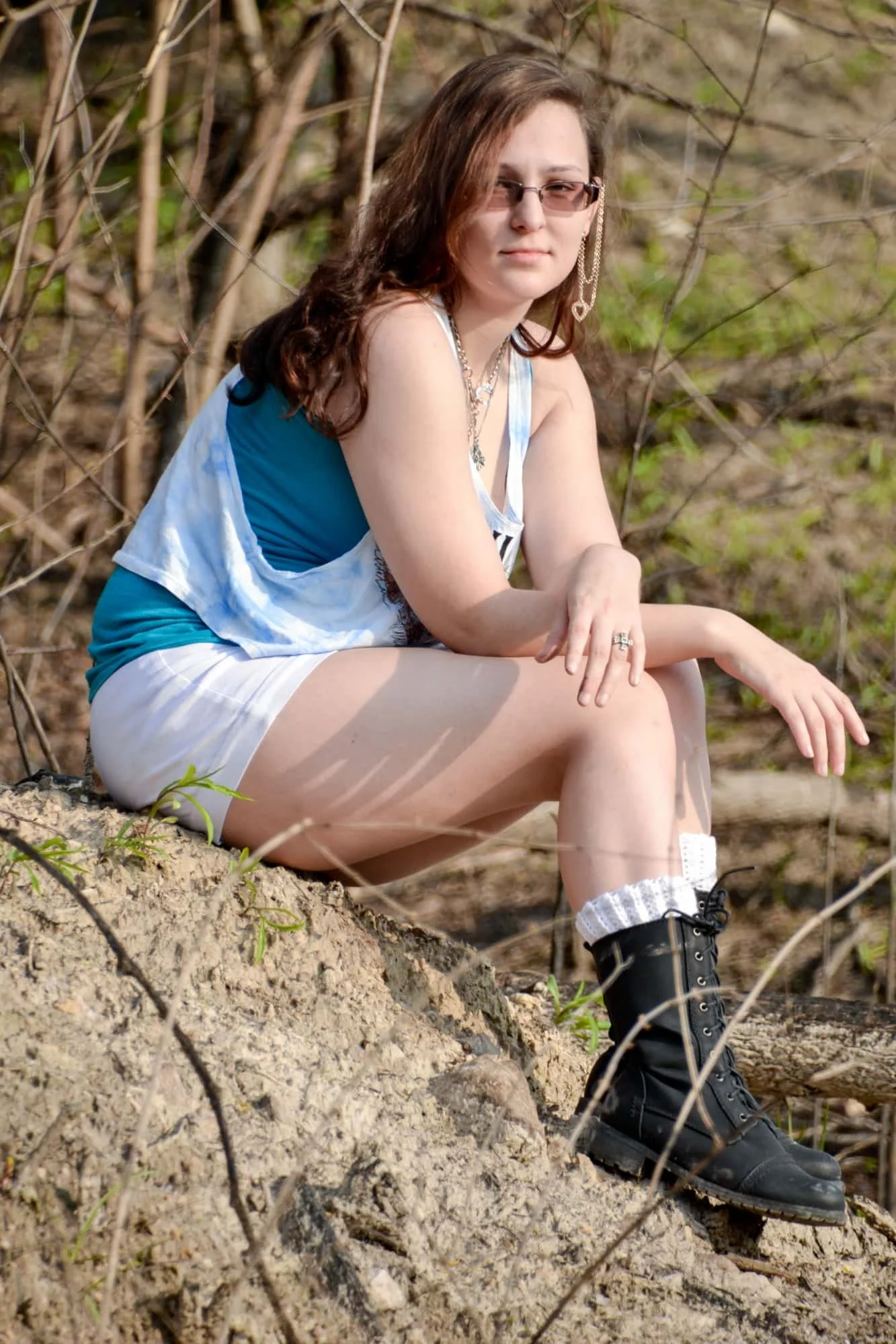 Senior girl wearing a white graphic tank, shorts, and black boots, sitting on a rock in the sunlight with her arms resting across her knees during a portrait session at Rock City in Valmeyer IL.