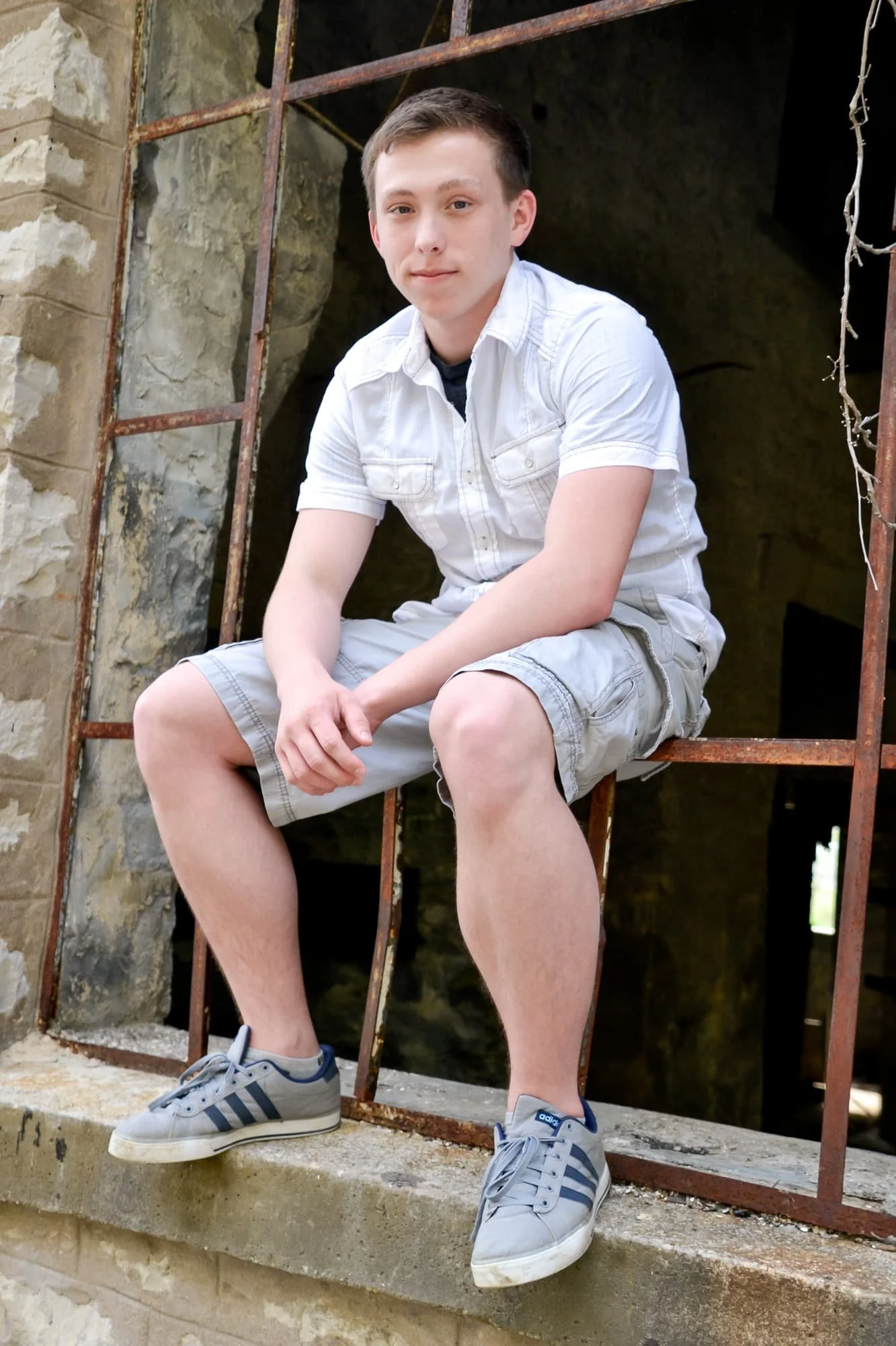 Senior photo of a guy wearing a white shirt, grey shorts, and grey shoes, sitting inside a rusted industrial window frame at Rock City in Valmeyer IL.