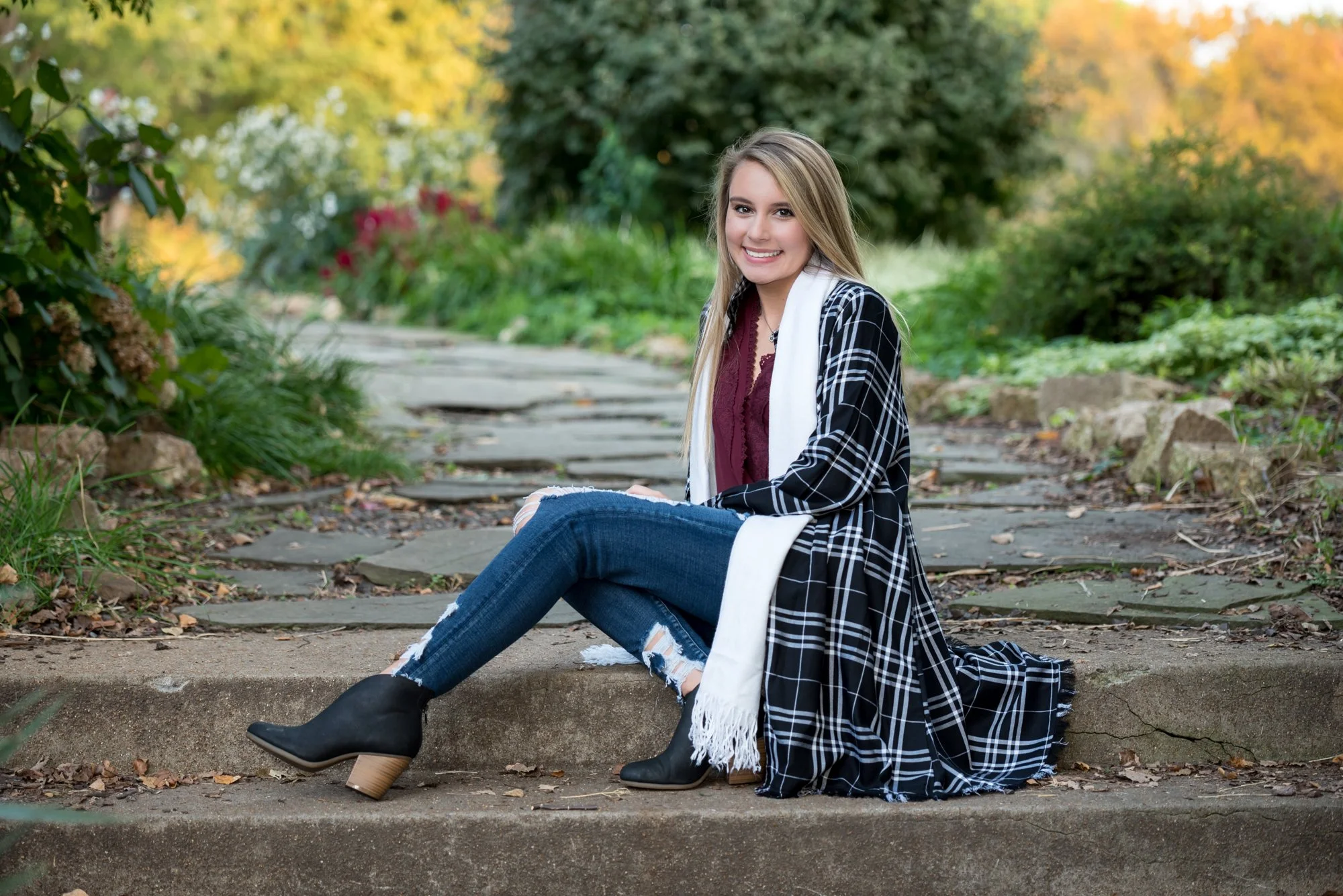 Boho style senior picture of a girl wearing a long black plaid cardigan, maroon shirt, ripped blue jeans, and short black boots, sitting across historic stone steps with a sunlit fall pathway and autumn trees behind her.