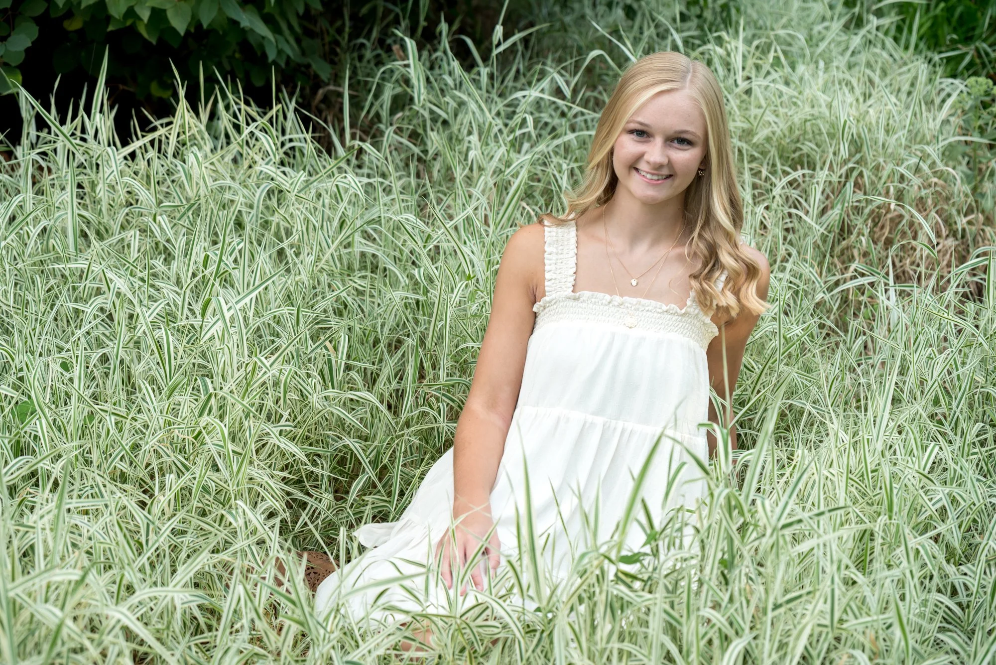 Whimsical senior photo of a girl in an airy white dress, sitting peacefully nestled in the tall green grasses of a sunlit park meadow.