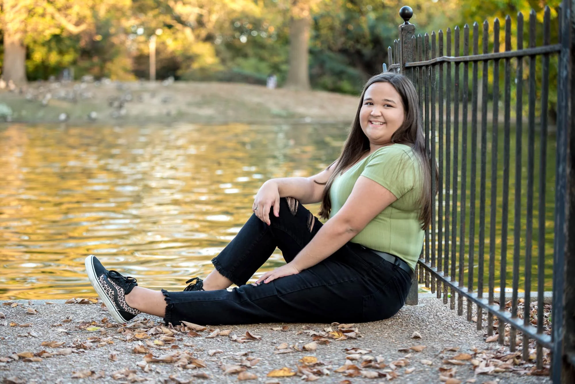 Beautiful autumn senior photo of a girl wearing a green t-shirt, black pants, and leopard print shoes, sitting next to a park pond and leaning back against a black wrought iron fence with peak fall foliage in the background at Lafayette Park.