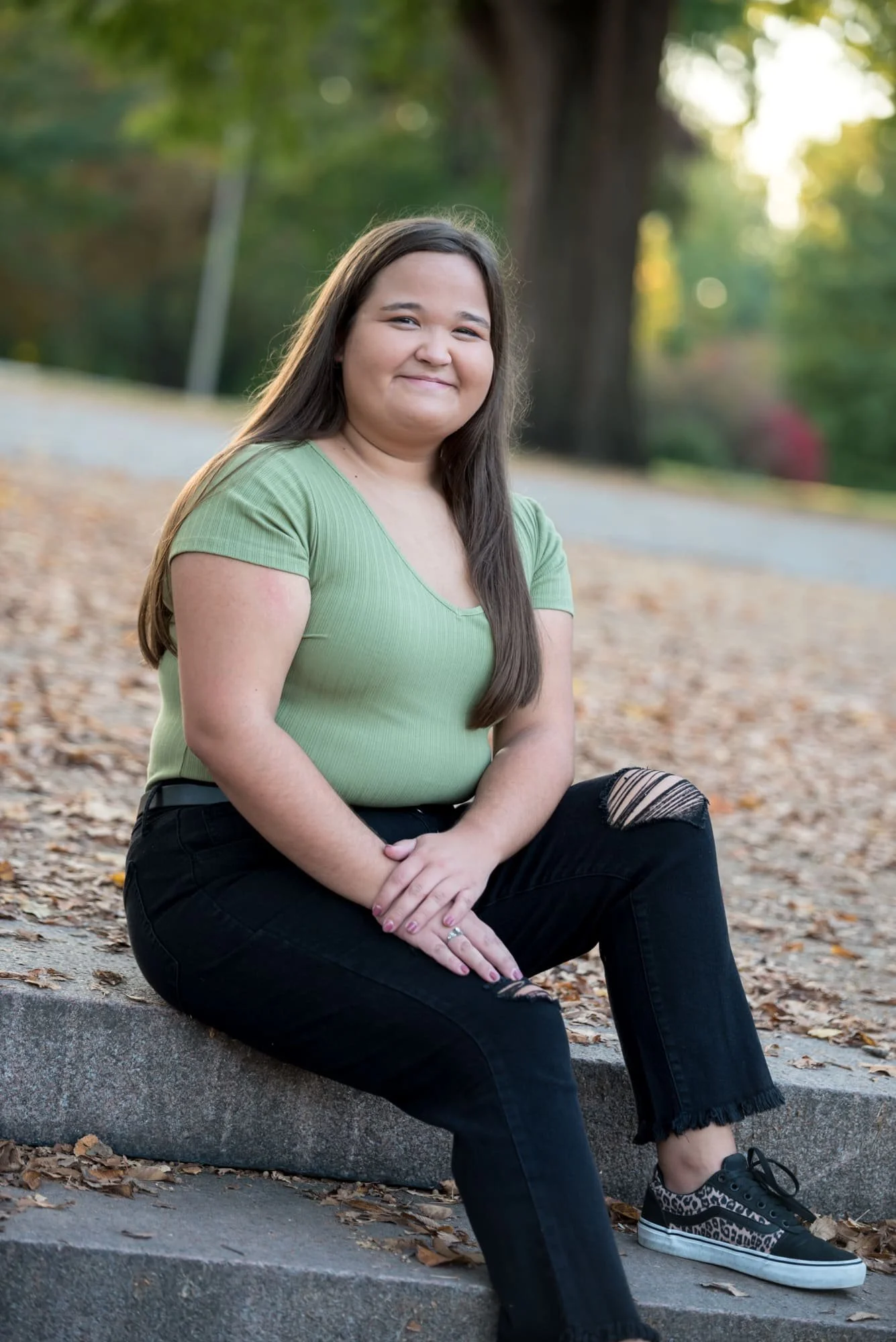 Classic fall senior picture of a girl wearing a green shirt and black ripped pants with leopard print shoes, sitting on stone park steps covered in bright orange autumn leaves in St. Louis, MO.