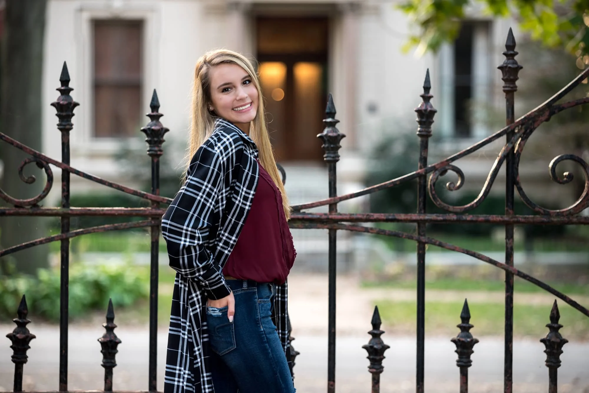 Beautiful autumn senior photo of a girl looking back over her shoulder, wearing a black and white plaid cardigan, maroon top, and blue jeans in front of a historic St. Louis home and wrought iron fence at Lafayette Park.