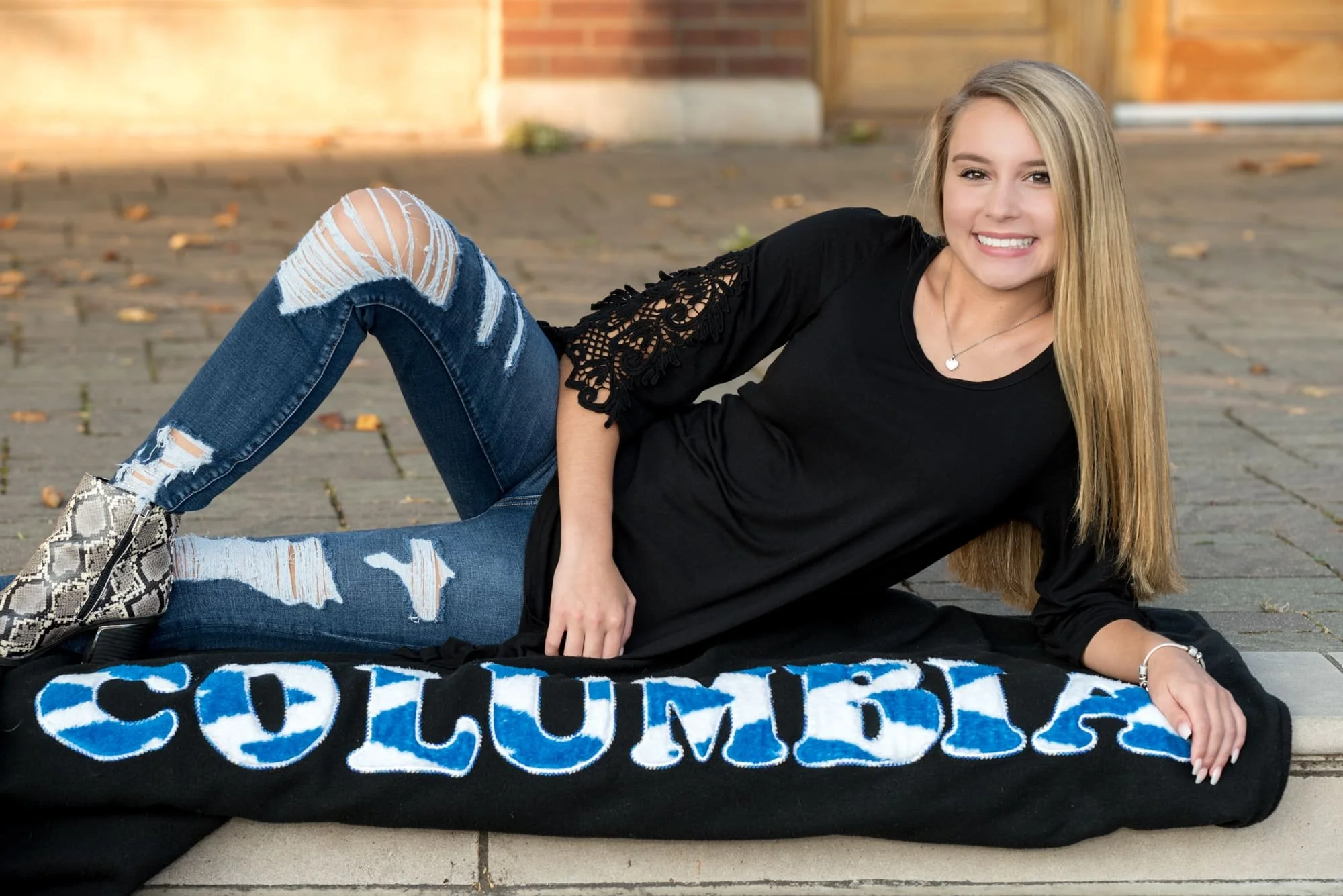 High school pride graduation photo featuring a girl wearing a white long-sleeve shirt, blue jeans, and stylish snakeskin boots, lounging across stone steps on a blue and white Columbia High School blanket.
