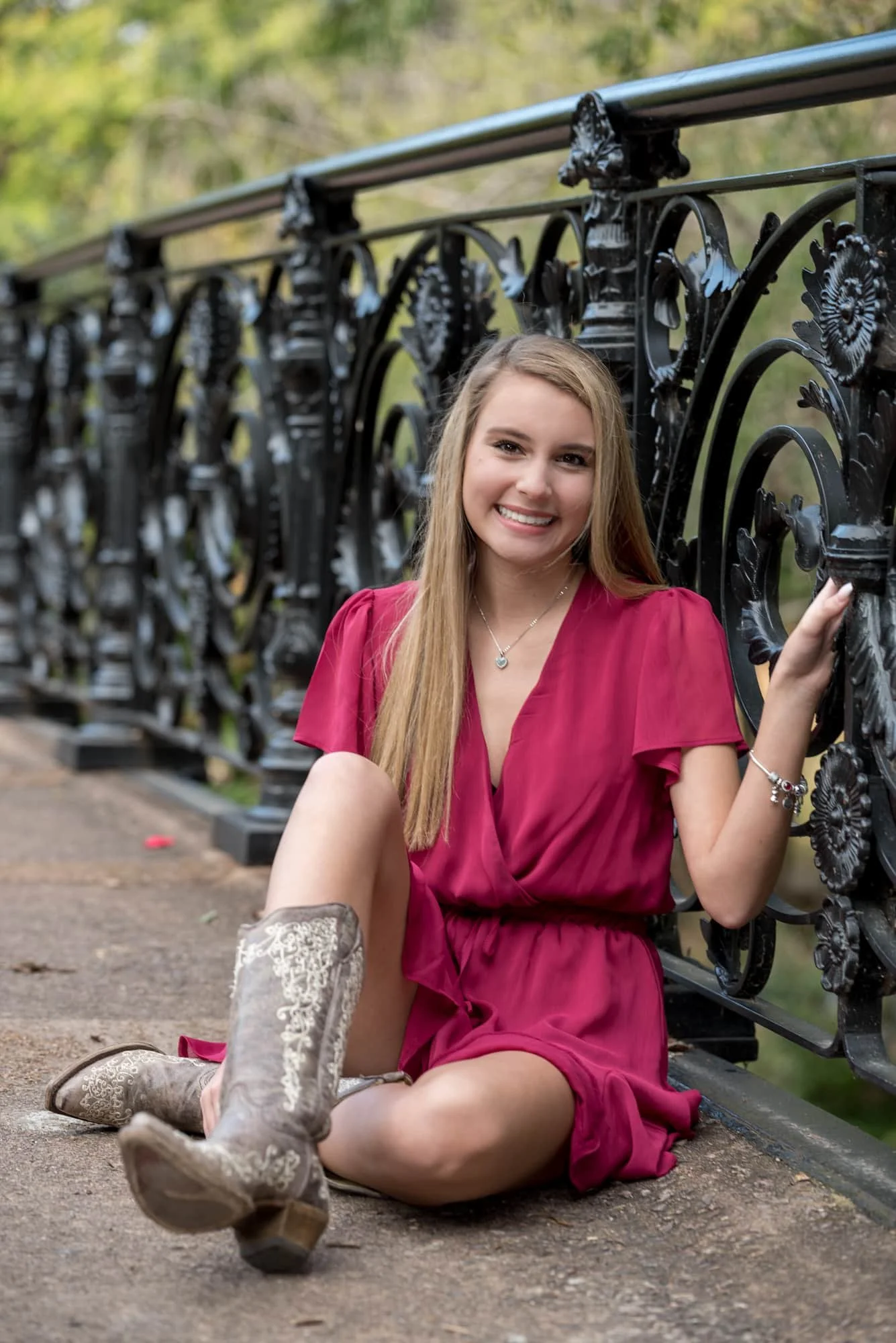 Country-style senior picture taken by a local St. Louis photographer, capturing a girl wearing a pink dress and cowboy boots while sitting on a historic park bridge with her hand on the black iron railing.