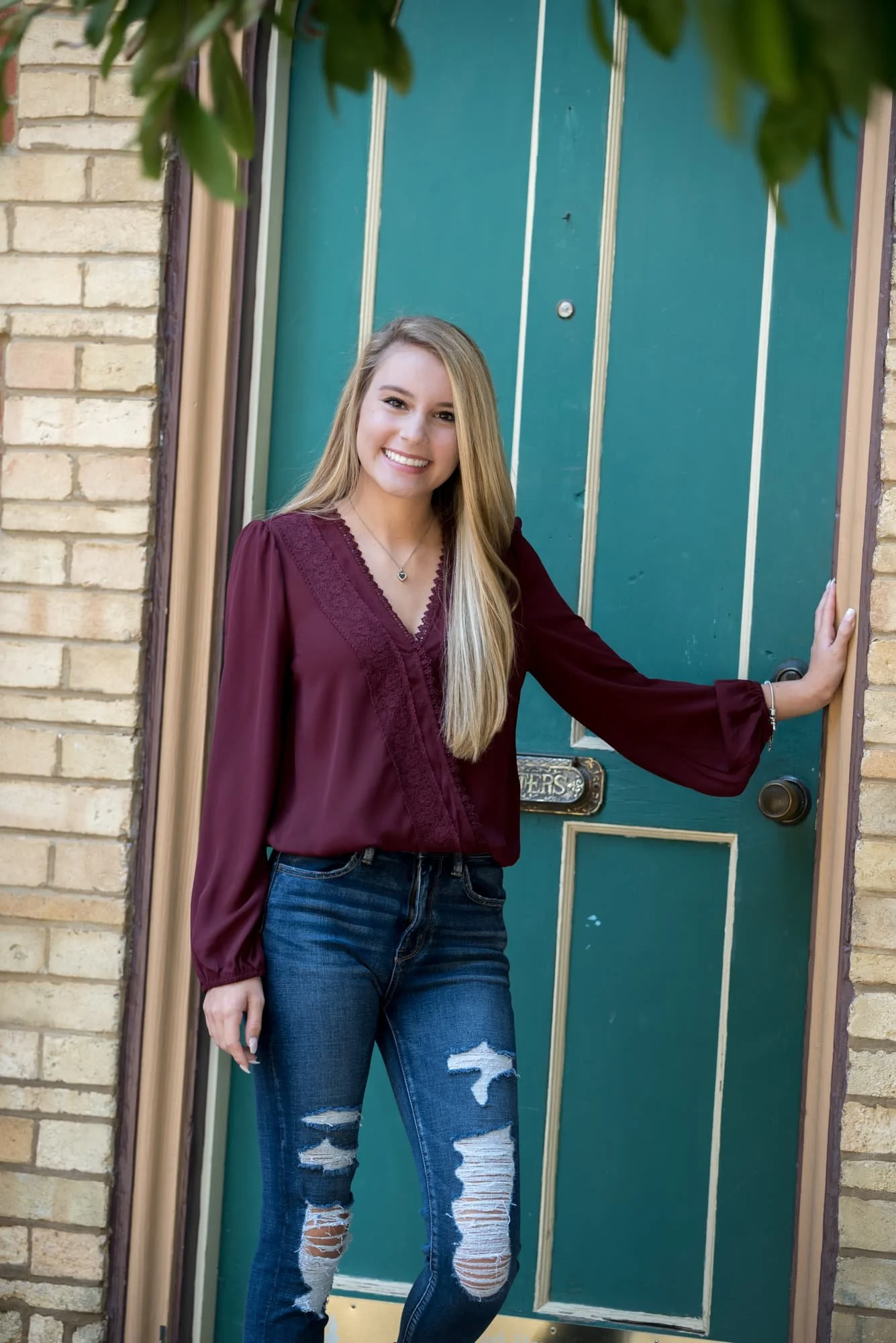 Rustic architectural senior photo of a girl wearing a maroon long-sleeve shirt and ripped blue jeans, standing in the doorway of a historic brick building framed by overhanging tree branches in St. Louis City.