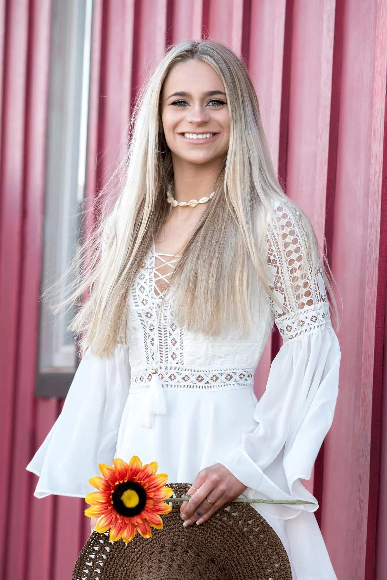 Whimsical country senior photo of a smiling girl wearing a white dress and a floppy brown hat, standing in front of a rustic barn while holding a bright orange flower.