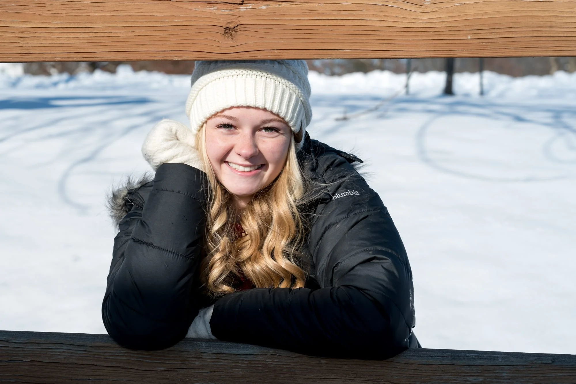Playful winter senior photo of a smiling girl wearing a white beanie and black coat, squatting between wooden fence railings in front of a deep, snow-covered field at Suson Park.