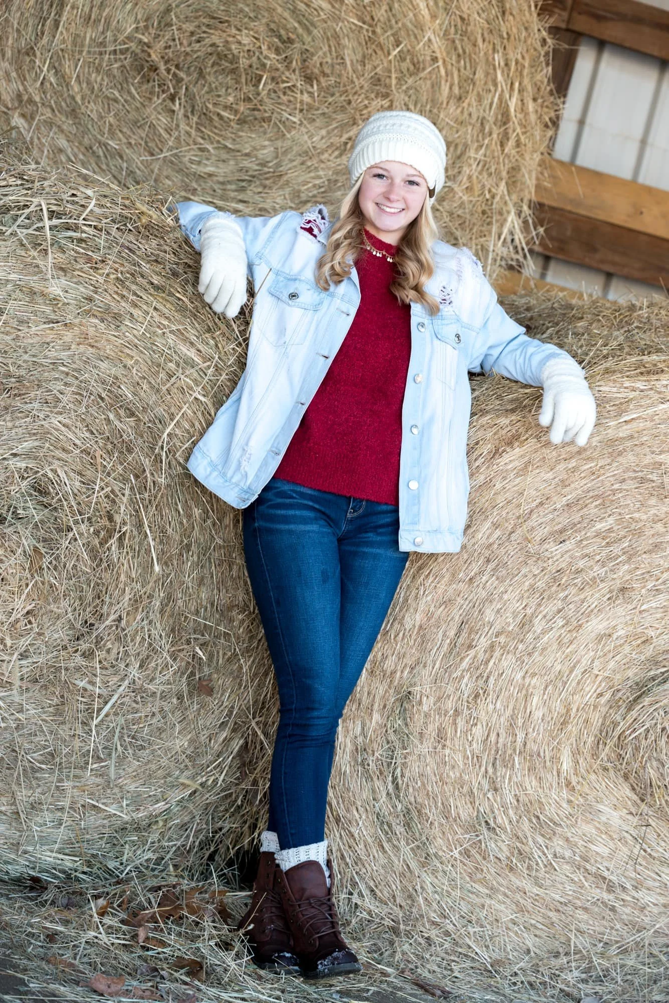 Cozy, rustic senior picture taken by a local photographer, featuring a girl wearing a red sweater and a white beanie, leaning her arms up against stacked hay bales inside a weathered barn.