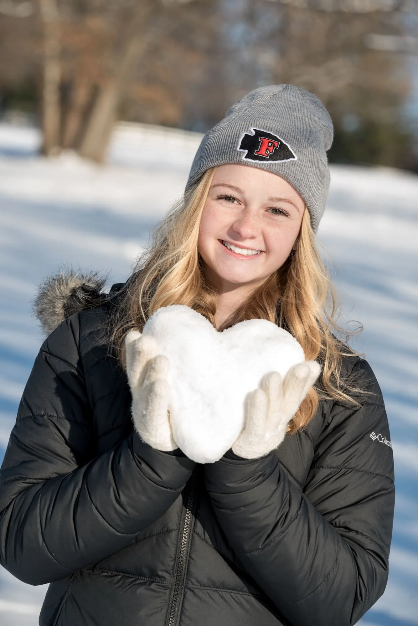 Classic wintry senior picture of a smiling girl wearing a black coat, white gloves, and a gray Fox High School beanie, holding up a heart shaped out of white snow against a blurred tree background.