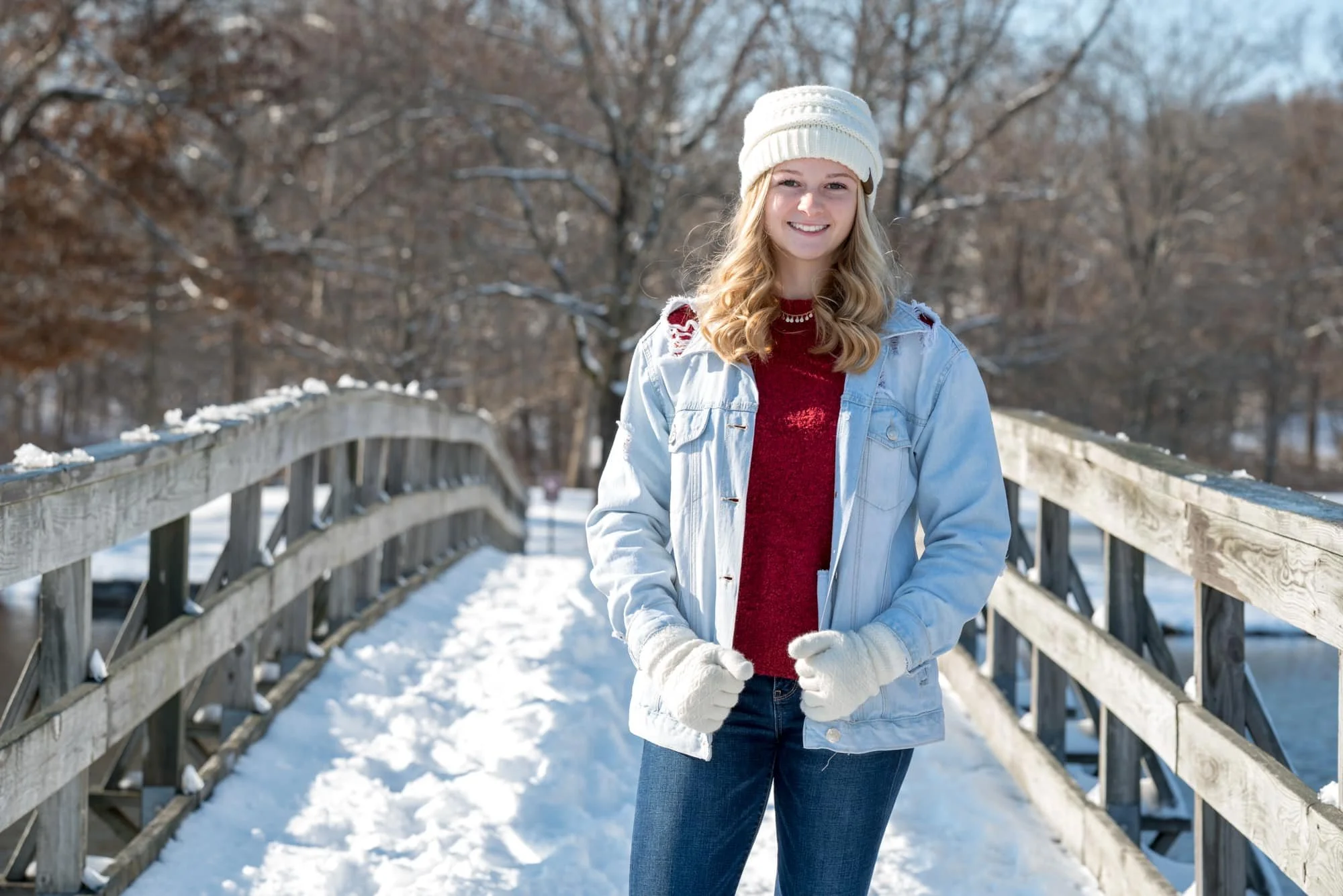 Winter senior photo of a girl wearing a light blue jean jacket and a red sweater, grabbing the bottom of her jacket while posing on a snow-covered wooden bridge over a lake in Arnold, Missouri.