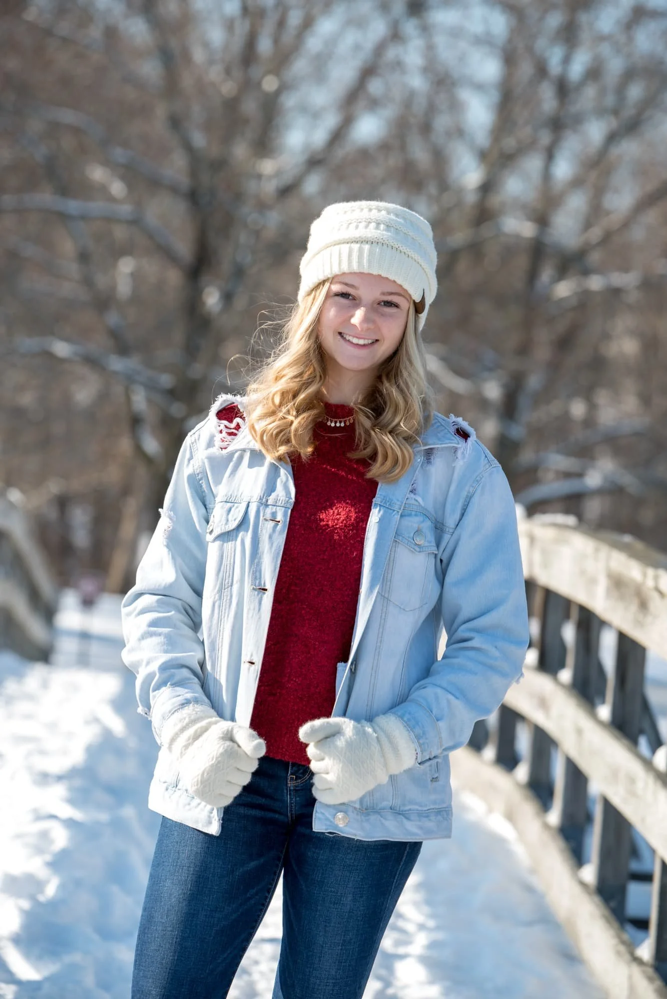 Senior girl standing on a wooden bridge in the winter snow at Suson Park, holding a light blue jean jacket while wearing a white beanie, red sweater, and white gloves.