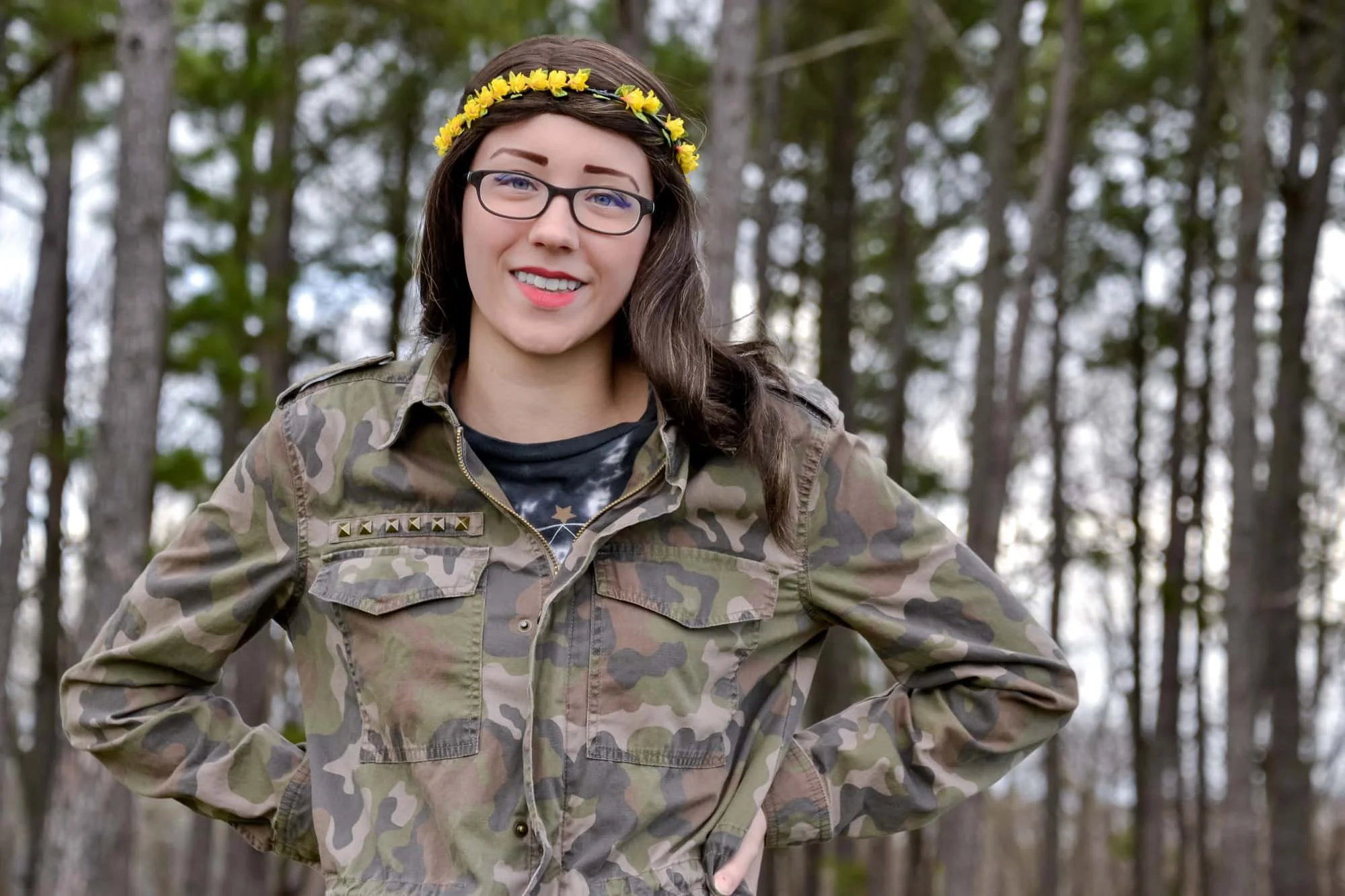 Senior girl wearing a camo jacket, glasses, and a yellow flower headband, standing with her hands on her hips in front of evergreen pine trees for a fall portrait at Konarcik Park in Waterloo IL.