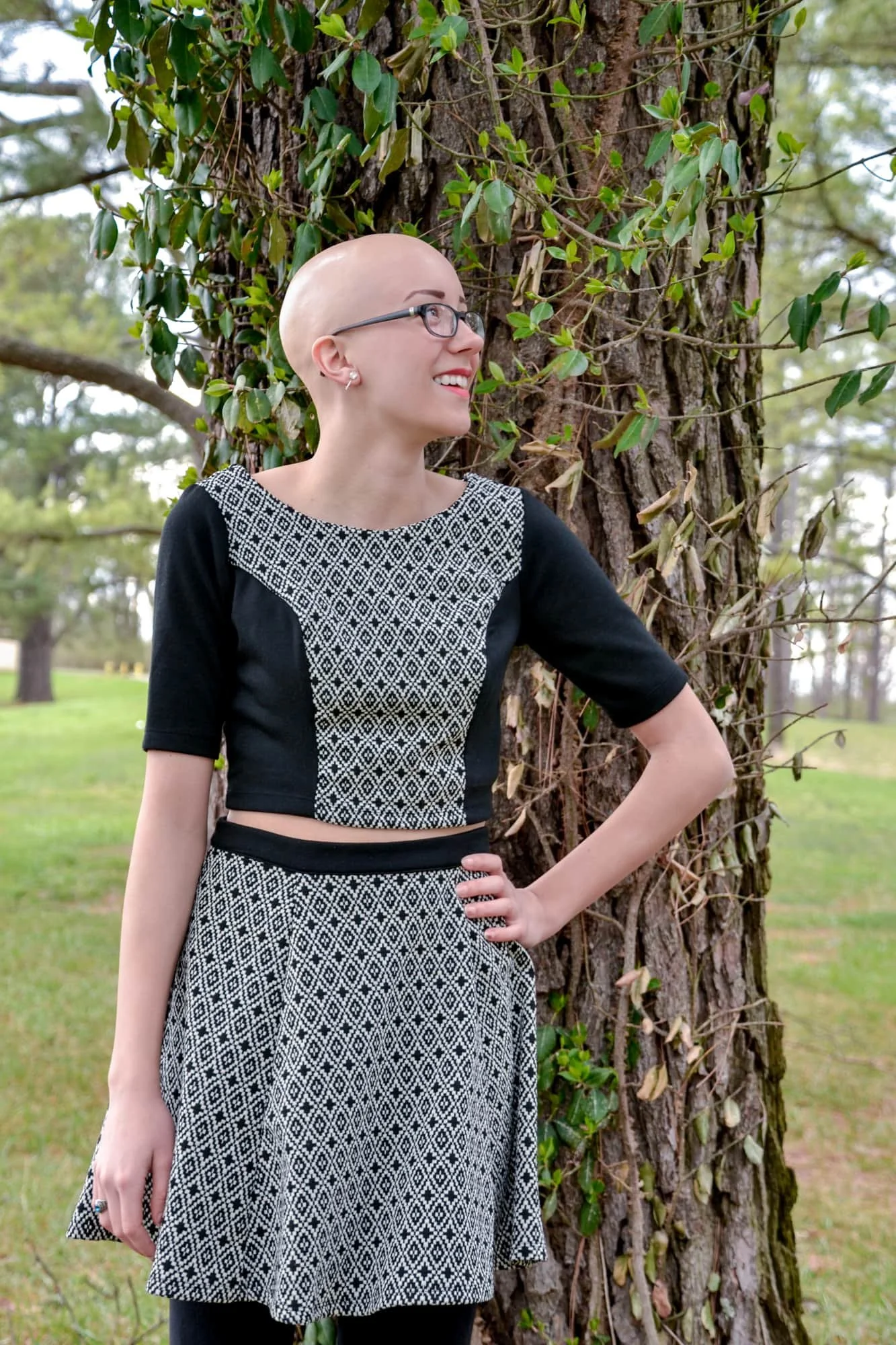 High school senior photo of a bald girl standing with her hand on her hip, looking off into the distance with a bright smile in front of lush spring trees in Monroe County.