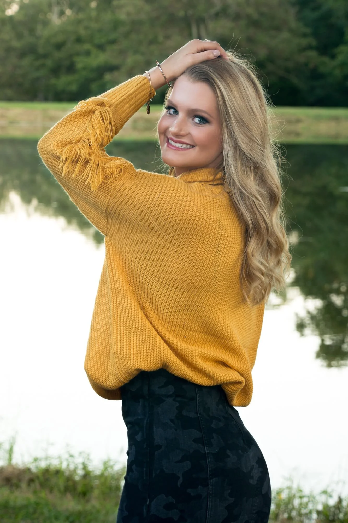 Senior photo of a girl wearing a cozy yellow sweater and a black camo skirt, standing sideways with her hand on her head in front of a peaceful lake at Konarcik Park in Waterloo IL.