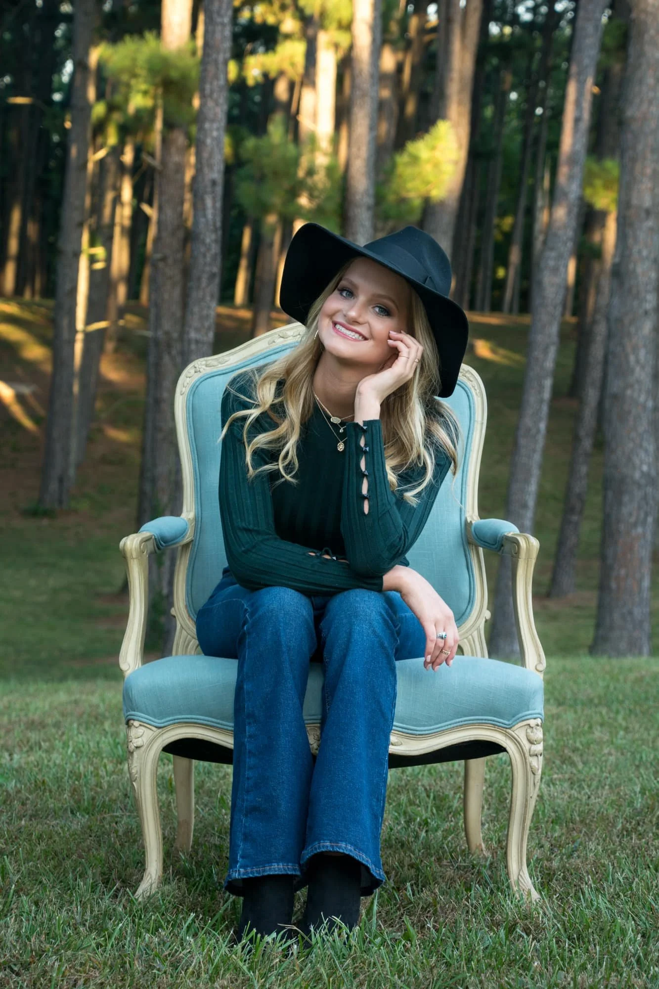 Senior picture of a girl with a gentle smile, wearing a black floppy hat and green long-sleeve shirt, sitting in a blue chair in front of a dense pine forest at Konarcik Park.