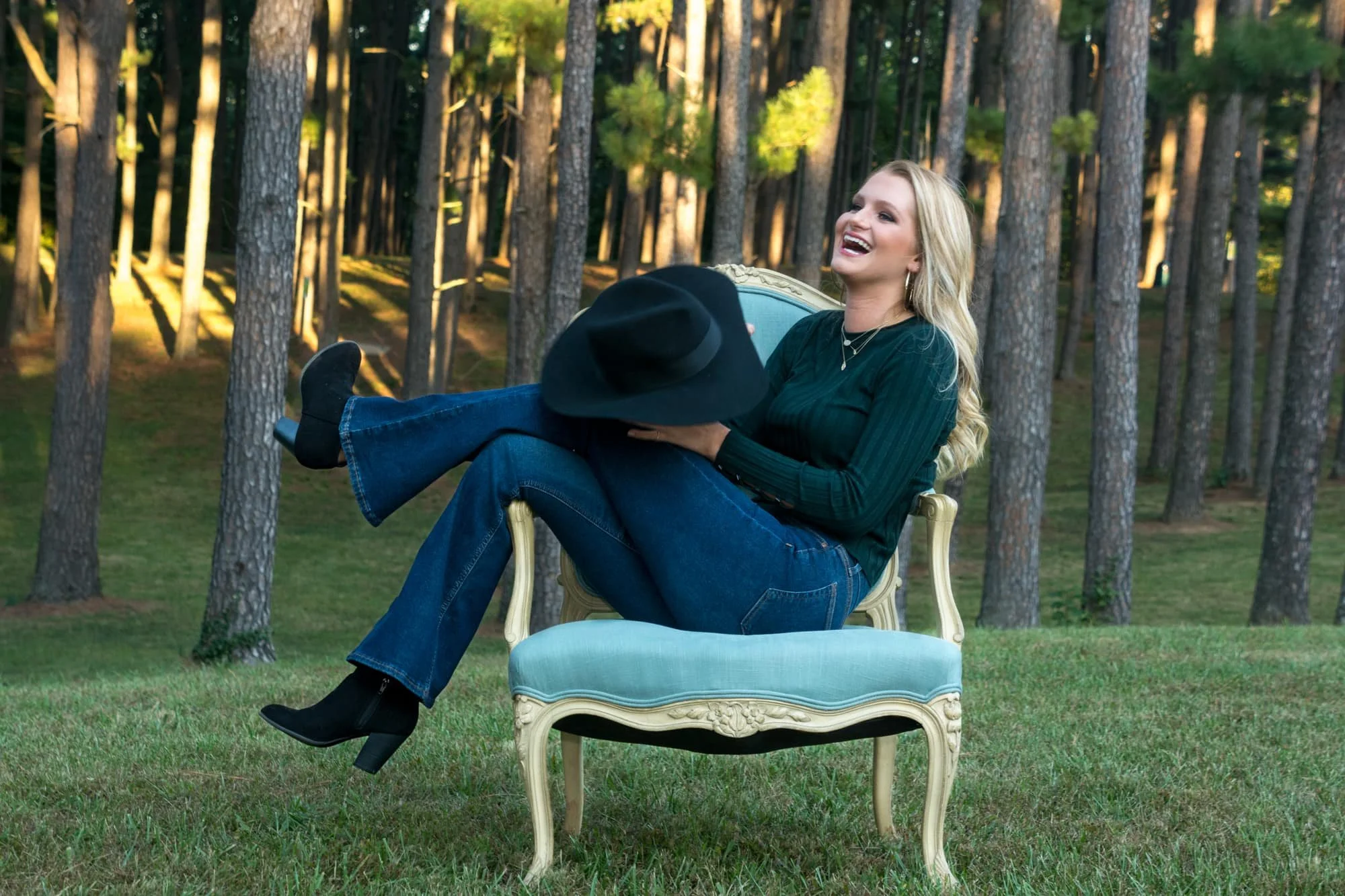 Candid senior picture of a girl laughing and holding her black floppy hat, sitting sideways in a blue chair in front of a beautiful pine forest backdrop.