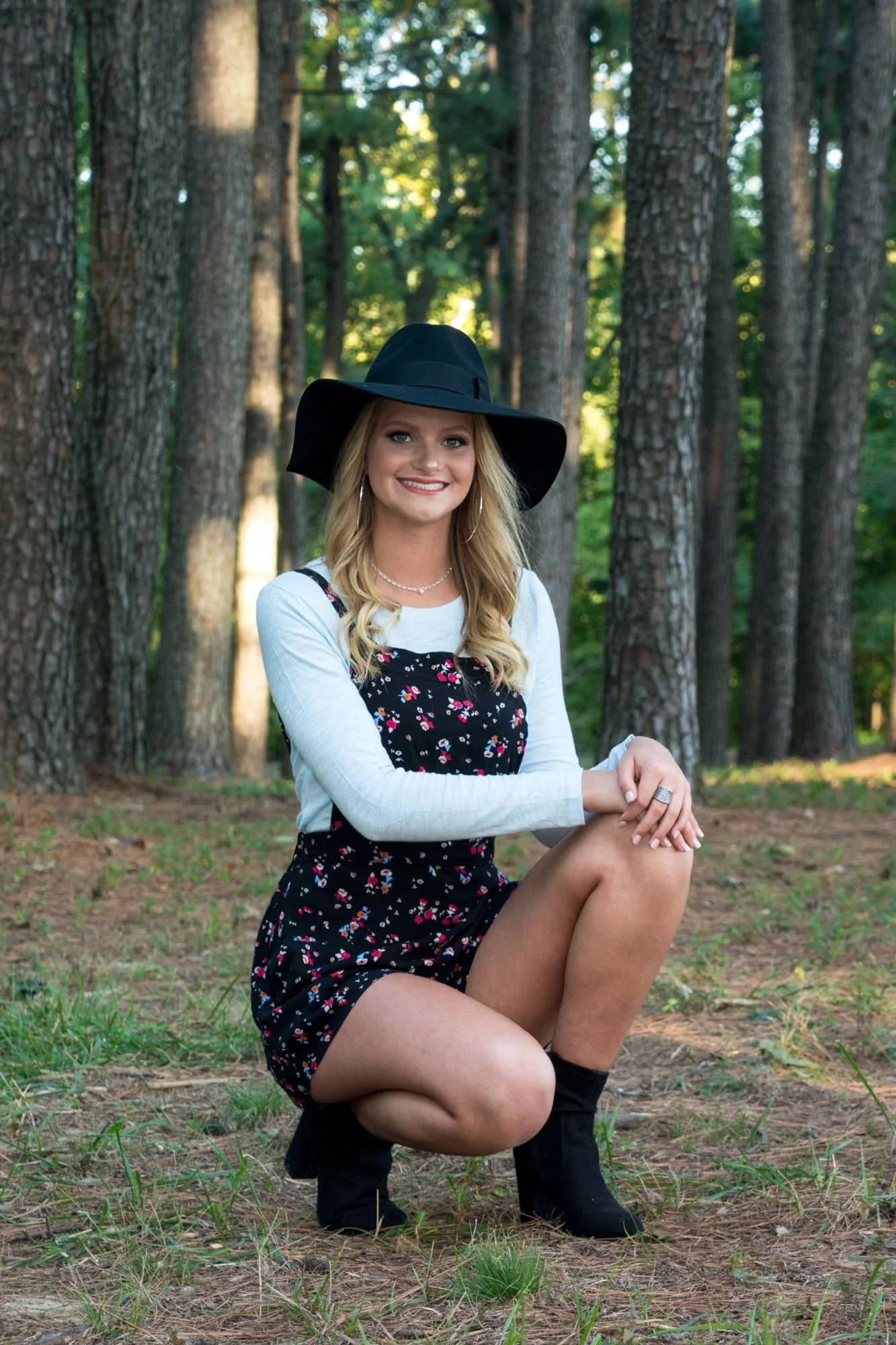 Alternative senior portrait of a girl wearing a black floppy hat, floral dress, and short black boots, squatting down in the pine needles in front of majestic pine trees in Monroe County.