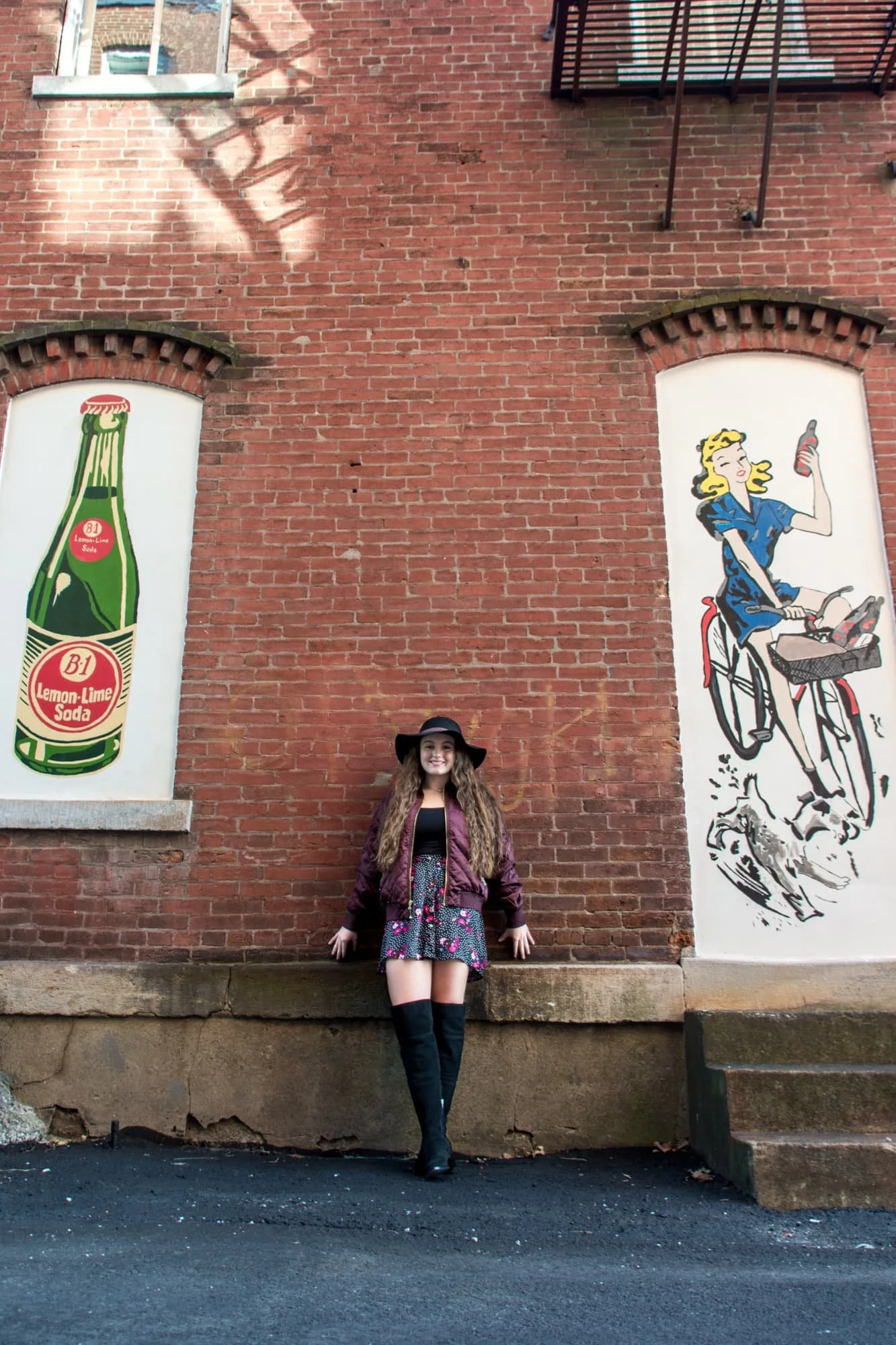 Senior picture of a girl leaning back against a brick building next to painted murals in downtown Waterloo IL, wearing a black floppy hat, black floral dress, purple jacket, and knee-high black boots.