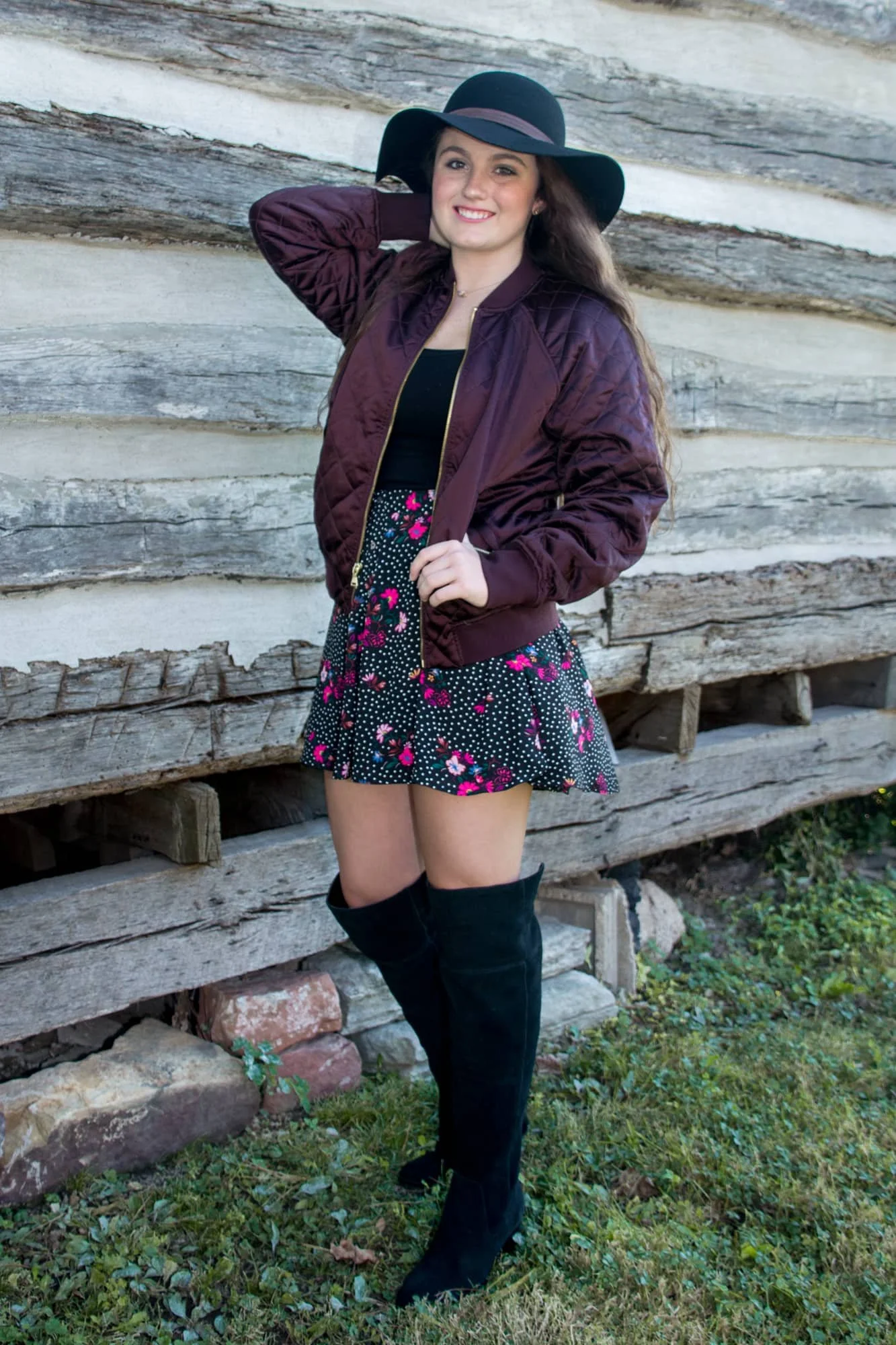 Senior picture of a girl standing next to a rustic log cabin, wearing a black floppy hat, black floral dress, purple jacket, and black knee-high boots with her hand in her hair.