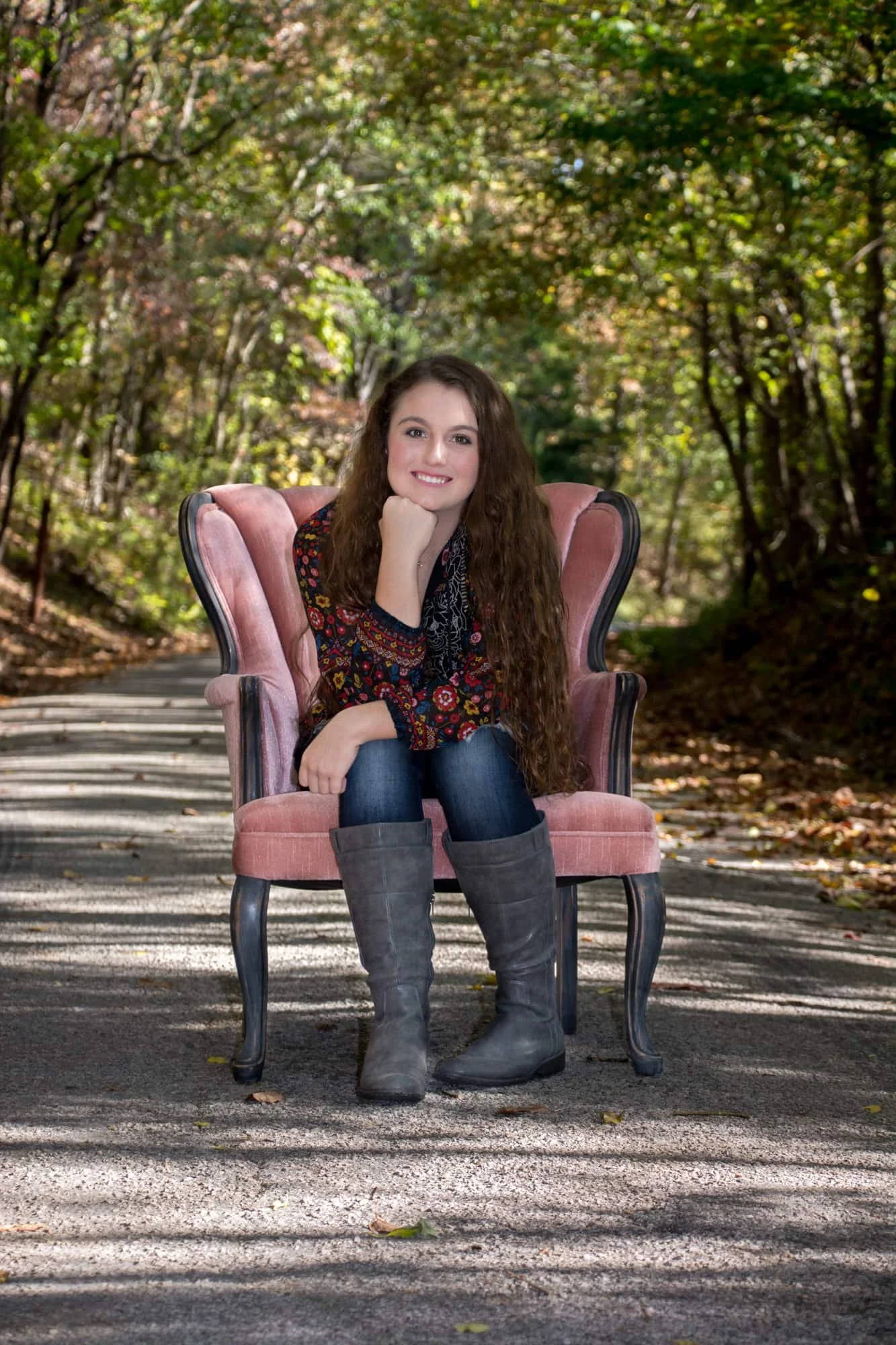 Senior picture of a girl sitting in a pink velvet chair in the middle of a fall roadway, wearing a multicolored black shirt, jeans, and grey boots while resting her chin on her hand.