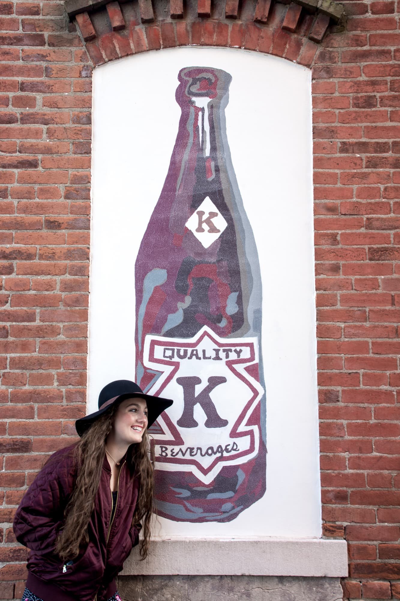 Senior girl laughing while standing next to a historic brick mural in Waterloo IL, wearing a black floppy hat and a purple jacket, taken by a Waterloo IL senior photographer.
