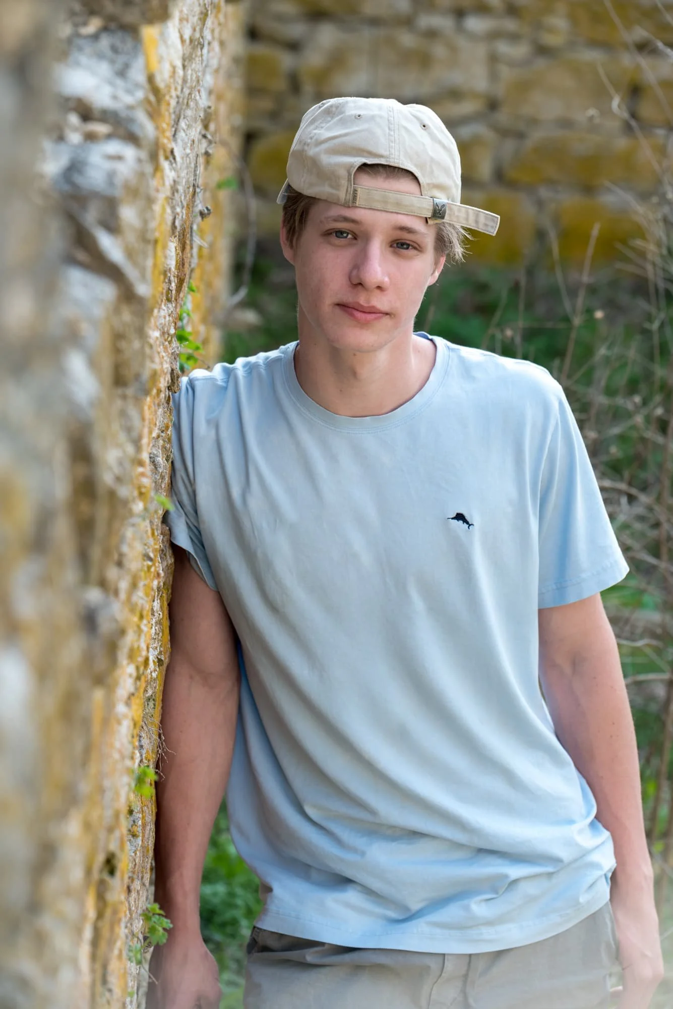 Serious senior picture featuring a boy in a light blue shirt, leaning confidently against a historic stone building covered in green moss in Columbia, IL.