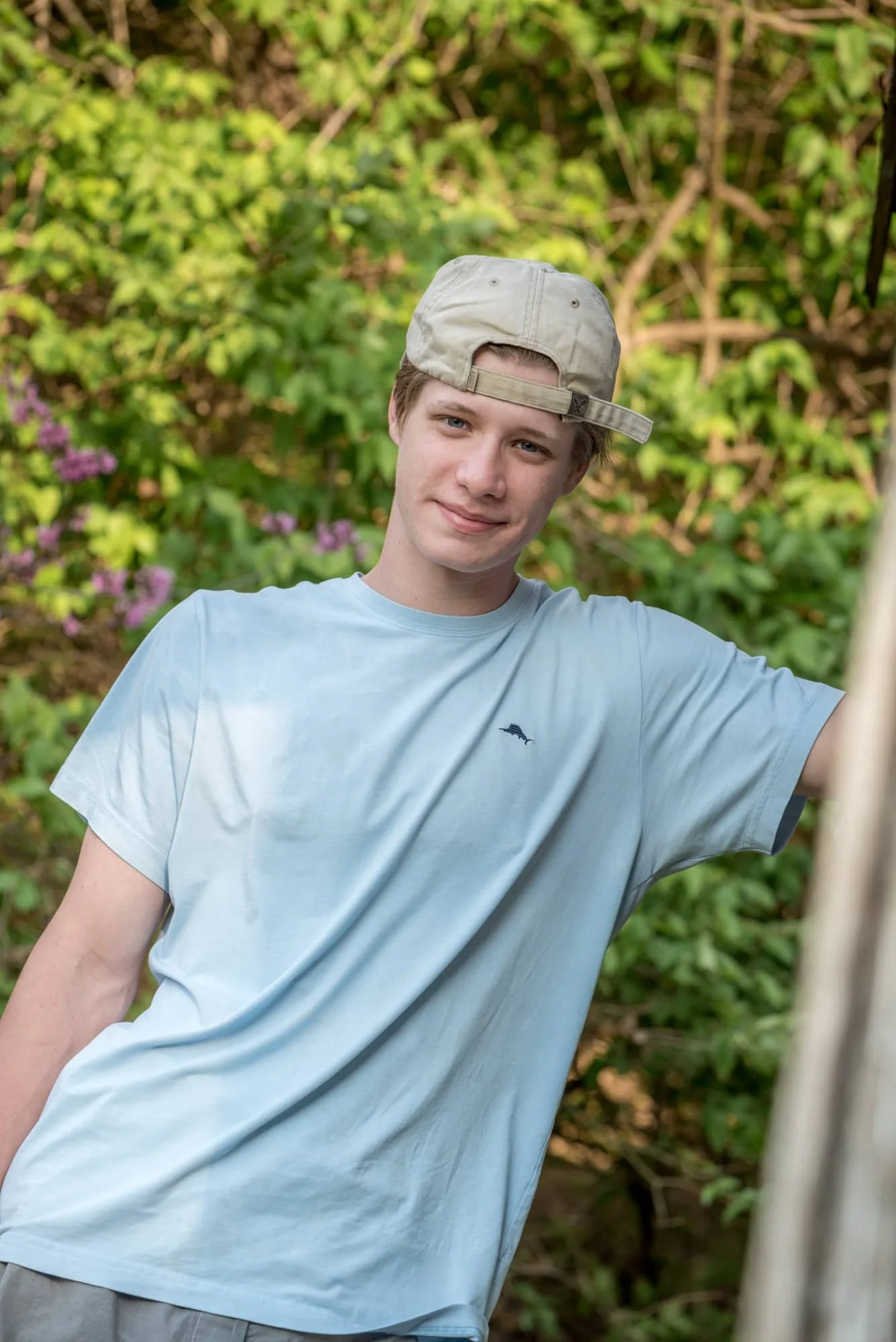 Natural senior picture taken by a local Columbia photographer, featuring a boy in a light blue shirt leaning against the side of a weathered wooden building with lush greenery behind him.