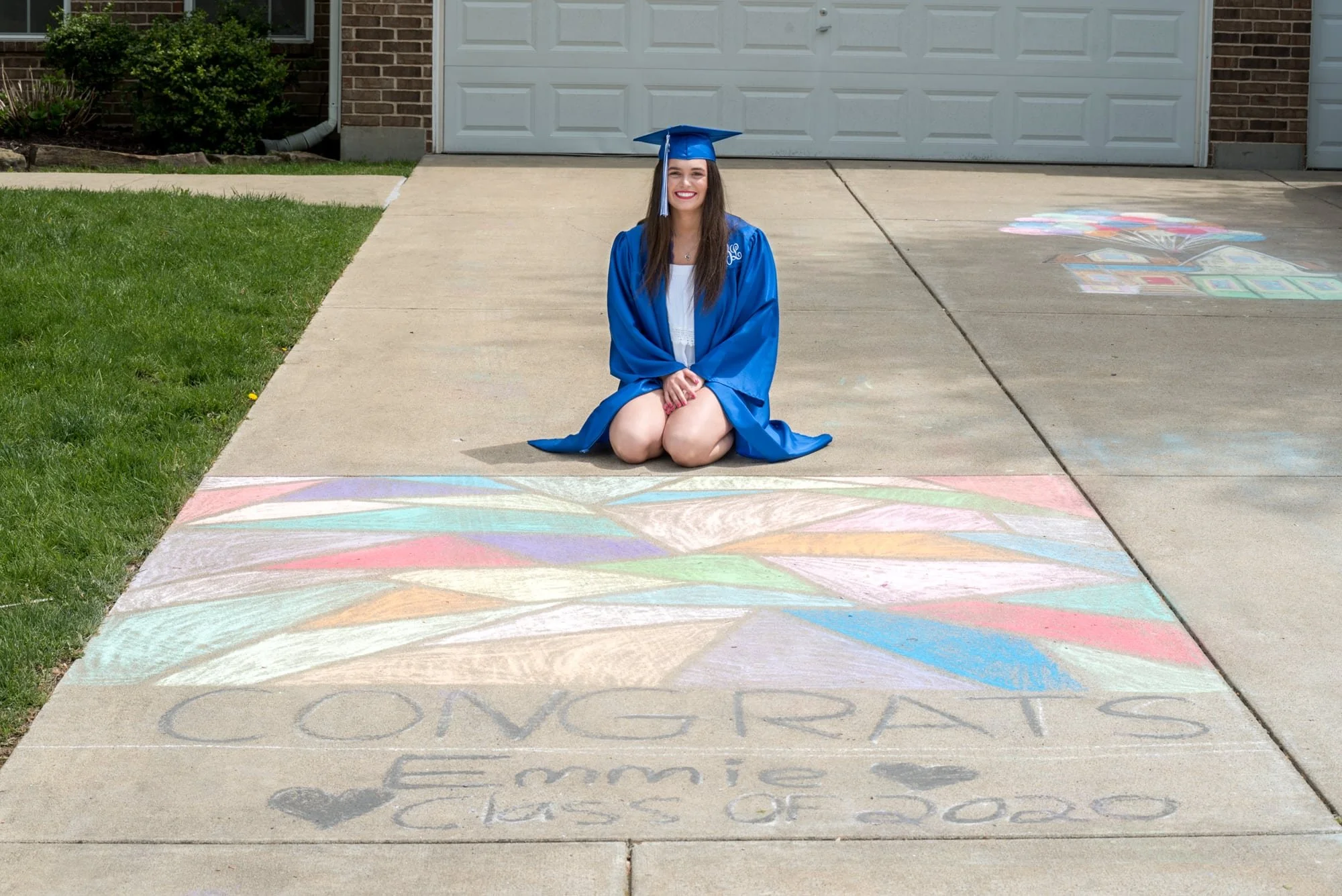 Fun graduation senior photo of a girl in her cap and gown sitting on her driveway behind a colorful chalk drawing that reads "Congrats Emmy Class of 2020," with her hands resting in her lap in Columbia, IL.