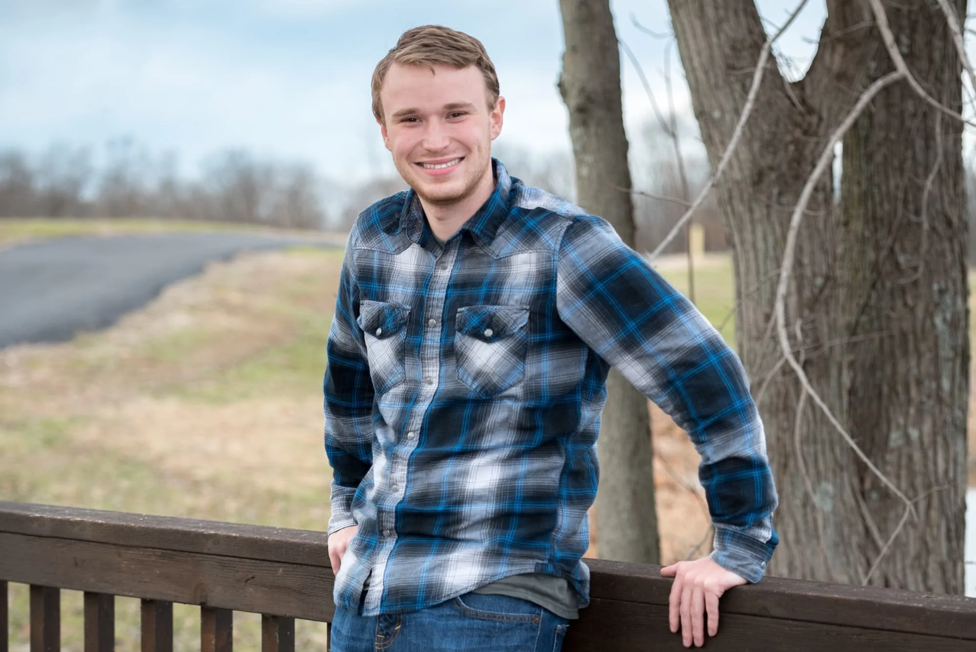 Senior guy standing against a wooden bridge on a trail during the winter, wearing jeans and a blue plaid shirt for his senior picture at Lakeview Park.