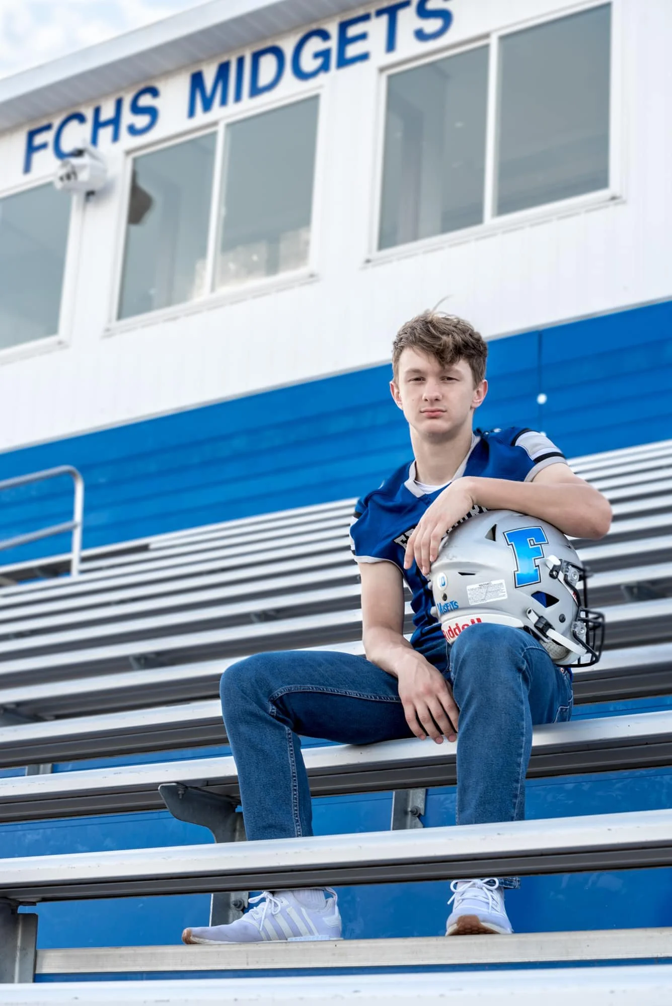 Senior guy athlete sitting on the bleachers at Freeburg High School for a football senior portrait session, wearing his jersey and jeans while holding his helmet with a serious expression.