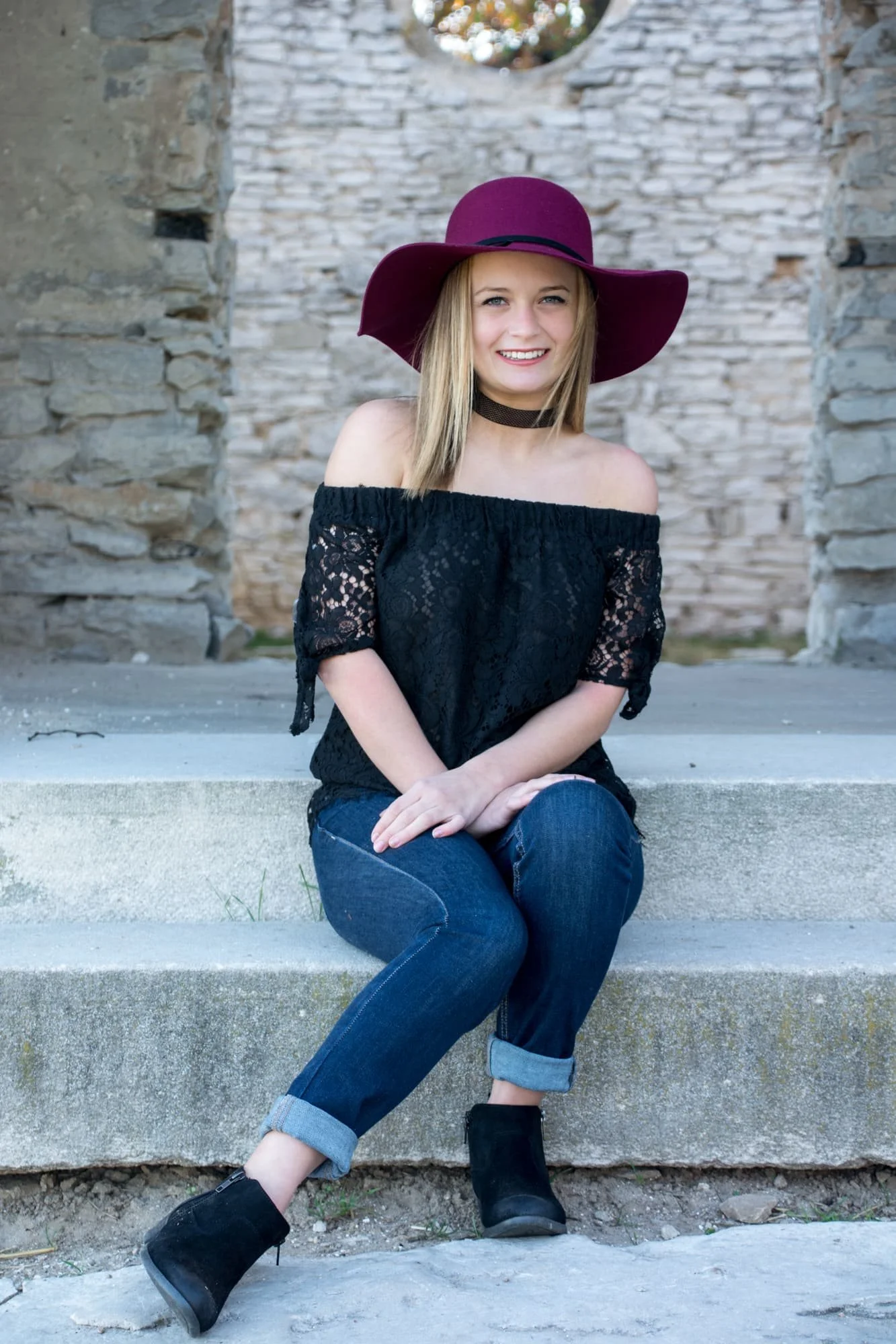 Senior picture of a girl sitting on the steps of Salem-Baum Church during fall with stone walls behind her, wearing an off-the-shoulder black lace top, blue jeans, short black boots, and a purple floppy hat with her legs to the side.