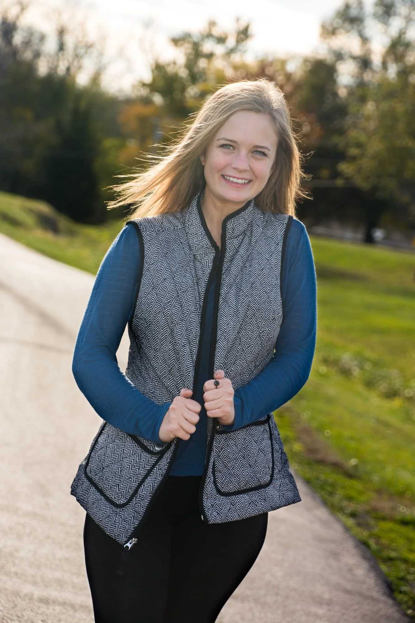 Senior girl standing on a pathway at Lakeview Park during a fall sunset, holding a black vest and wearing a teal long-sleeve shirt with black pants for her senior photo in Waterloo IL.